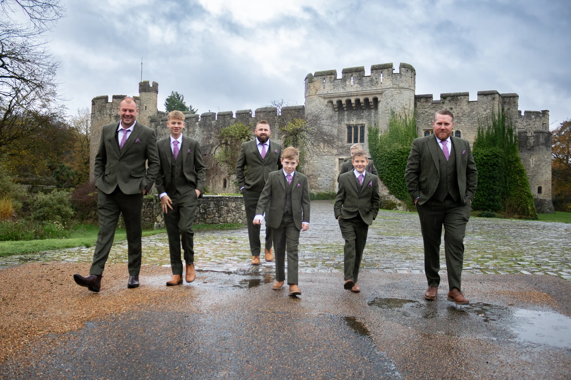 The groom and his groomsmen in matching tweed suits walk across a stone courtyard outside a historic castle.