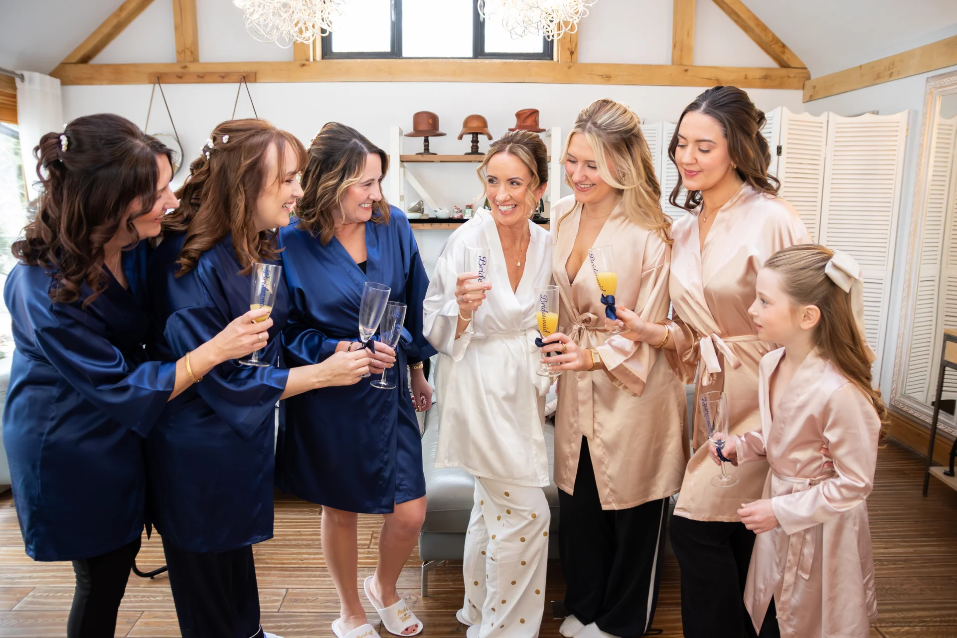A bride in a white robe and her bridesmaids in matching blue and pink silk robes raise glasses of champagne together during morning wedding preparations.