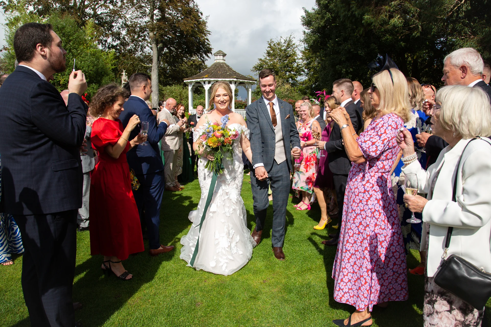 Newlyweds walk hand in hand through a cheering crowd of wedding guests holding drinks in a sunny garden with a bandstand in the background.