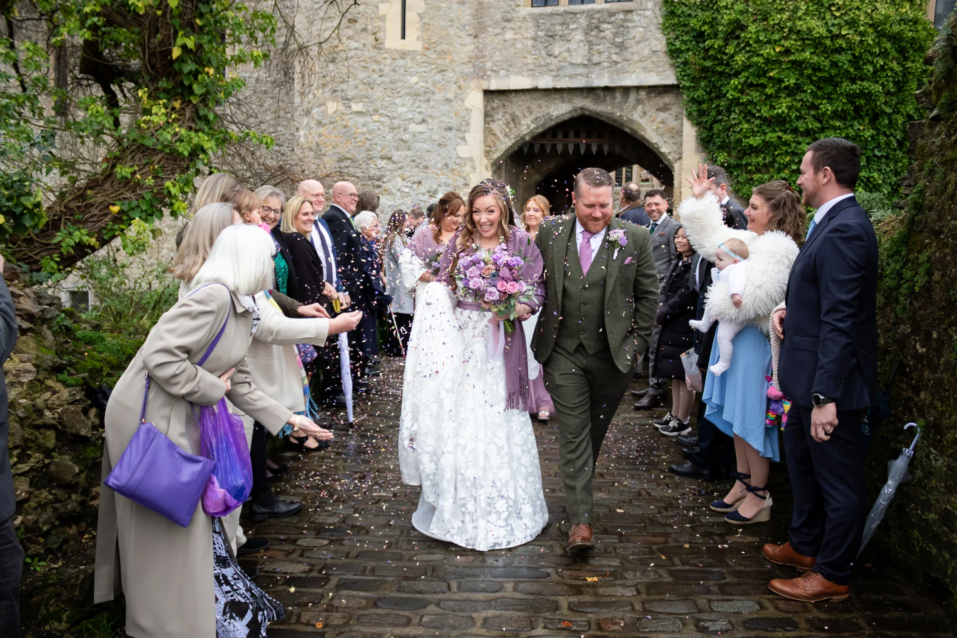 Newlyweds walk through a historic castle courtyard while smiling wedding guests shower them with colourful confetti.