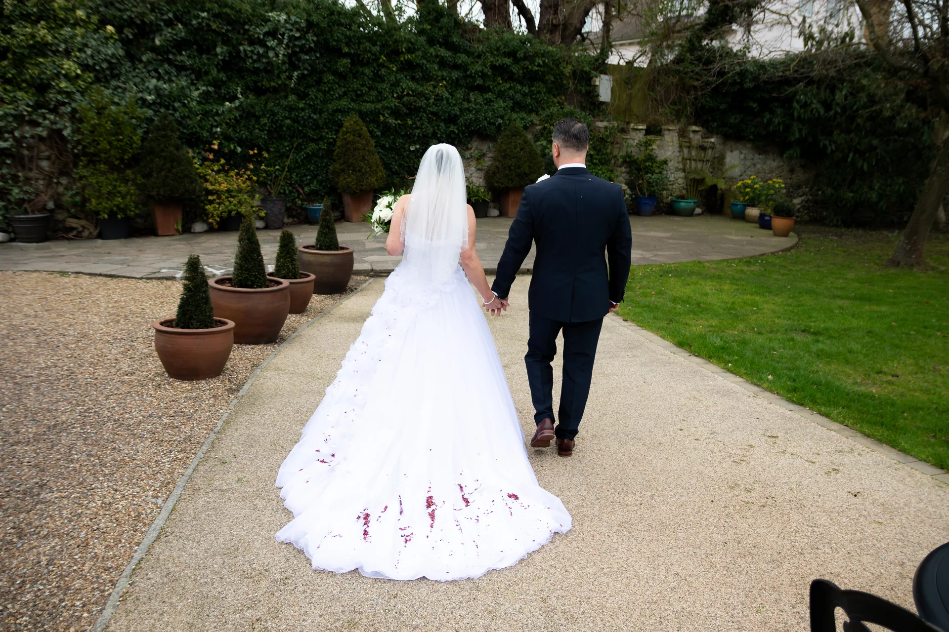 A newlywed couple walk hand in hand down a gravel path in a lush garden setting, viewed from behind with the bride's white gown featuring scattered petals.