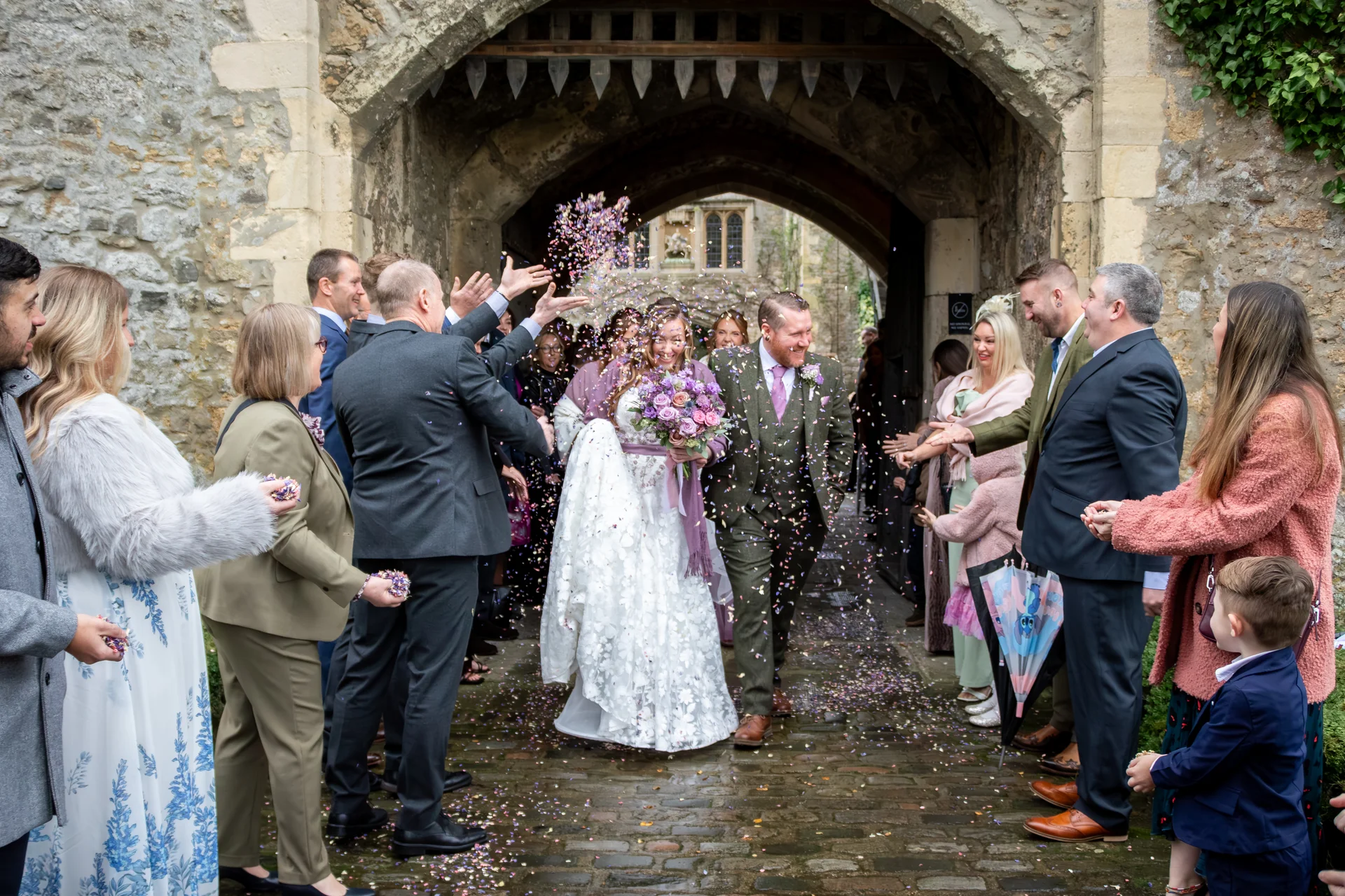 Newlyweds walk hand in hand through a historic stone archway as cheering wedding guests shower them with celebratory confetti.
