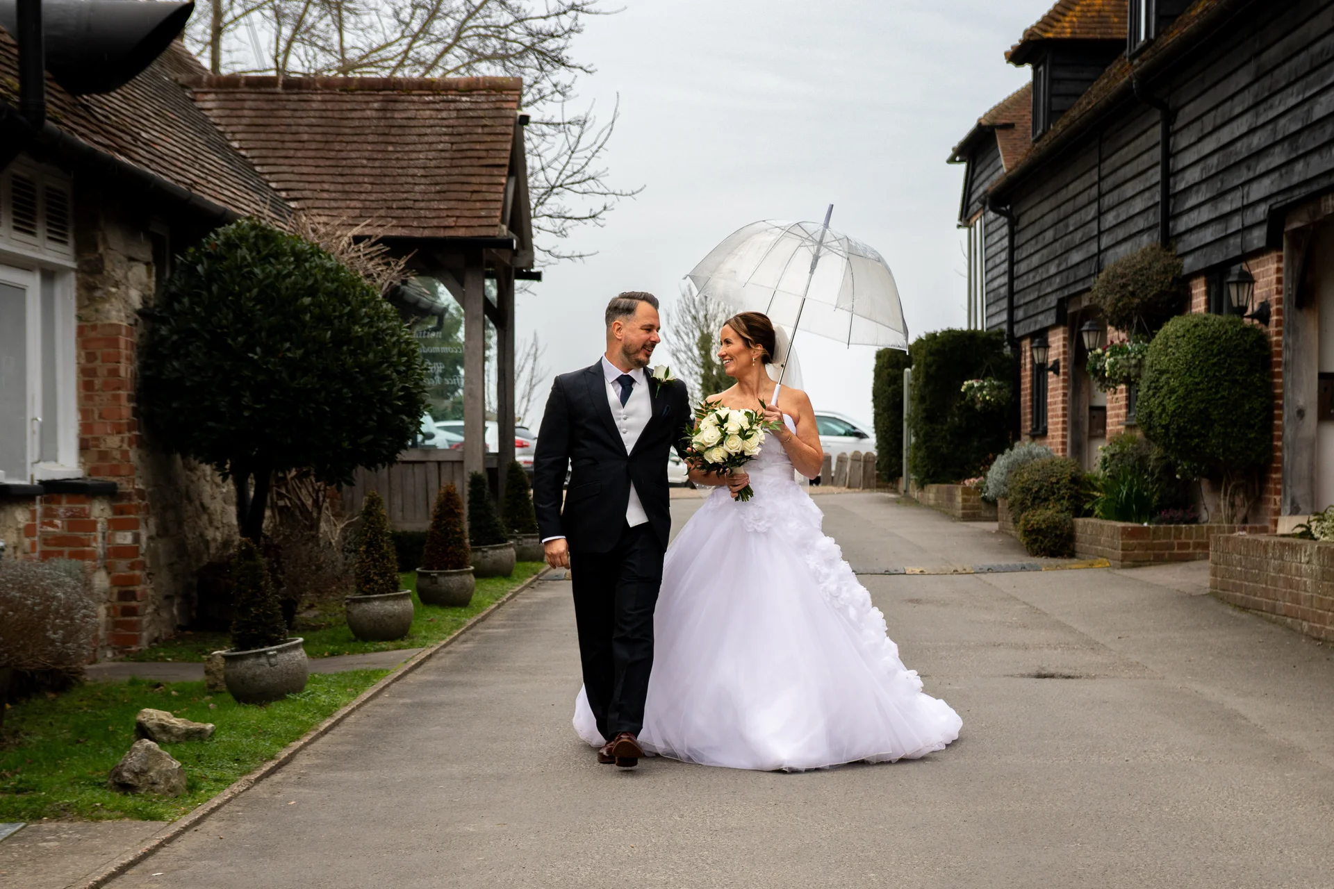 A newlywed couple walk hand in hand along a courtyard path between rustic buildings on their rainy wedding day, with the bride holding a clear umbrella and a white floral bouquet.