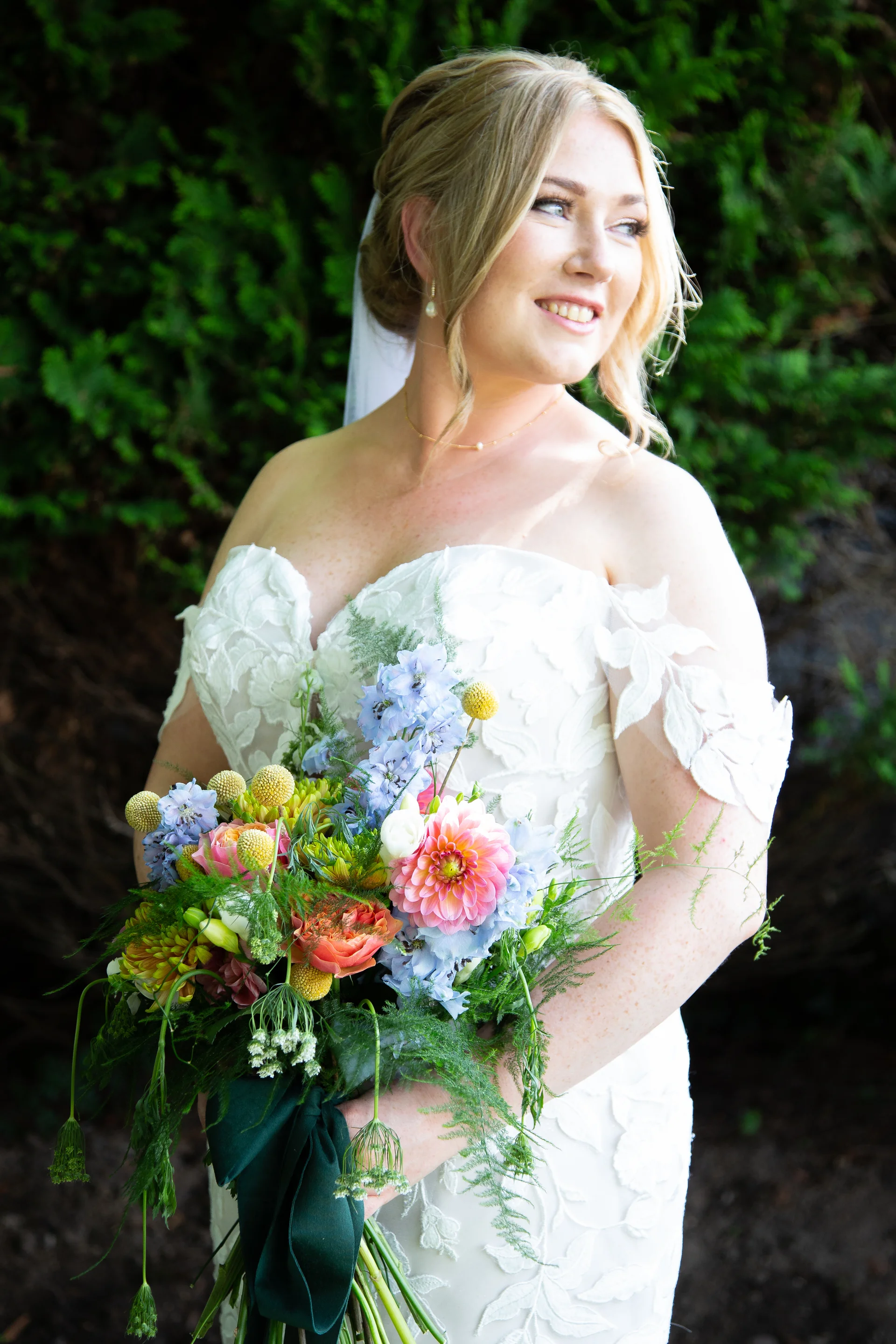 A smiling blonde bride wearing an off-the-shoulder gown with floral applique holds a colourful wild bouquet outdoors against a backdrop of dark foliage.