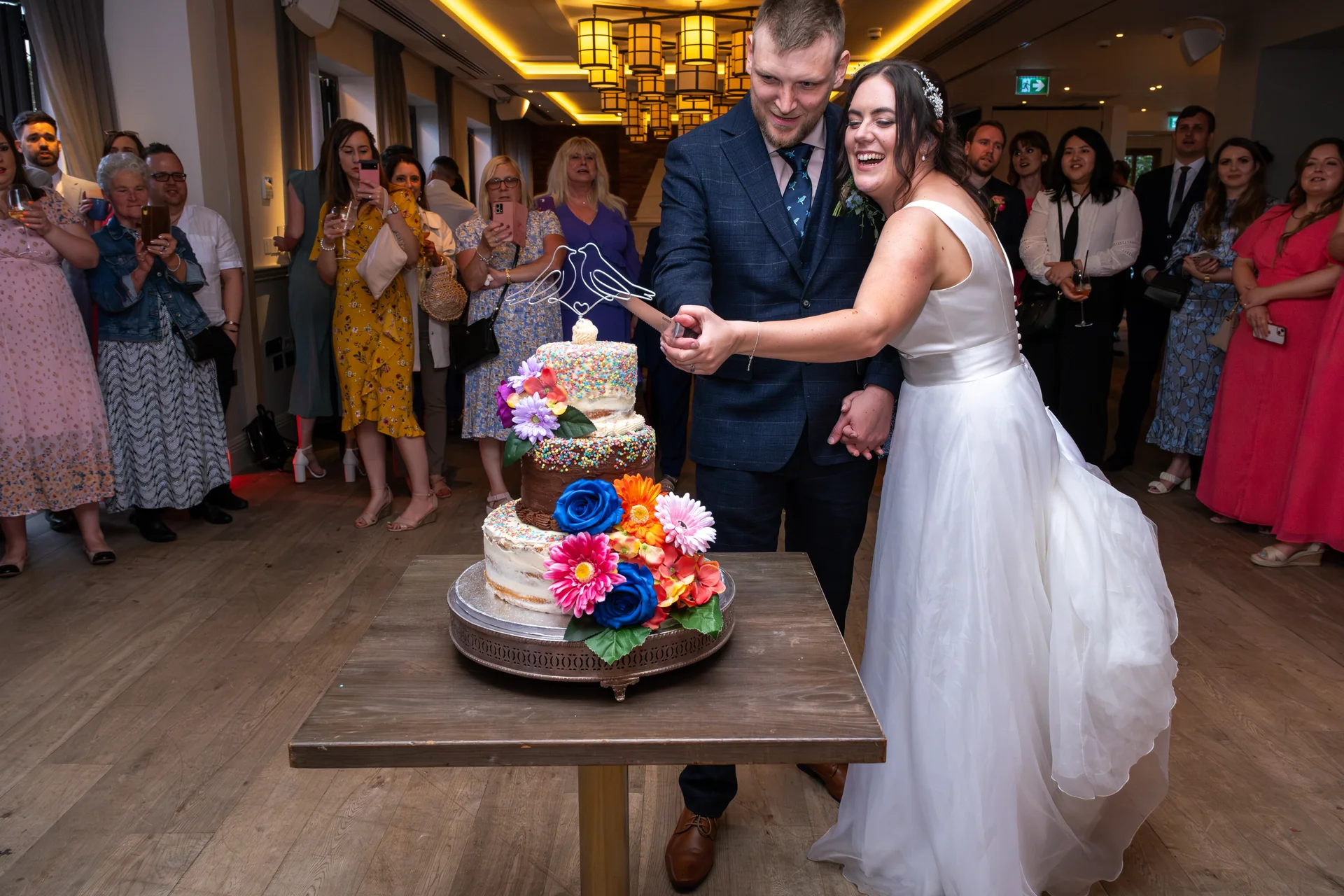 The bride and groom laugh joyfully while cutting a multi-tiered sprinkle-covered wedding cake decorated with bright vibrant flowers, surrounded by watching guests inside a hotel venue.