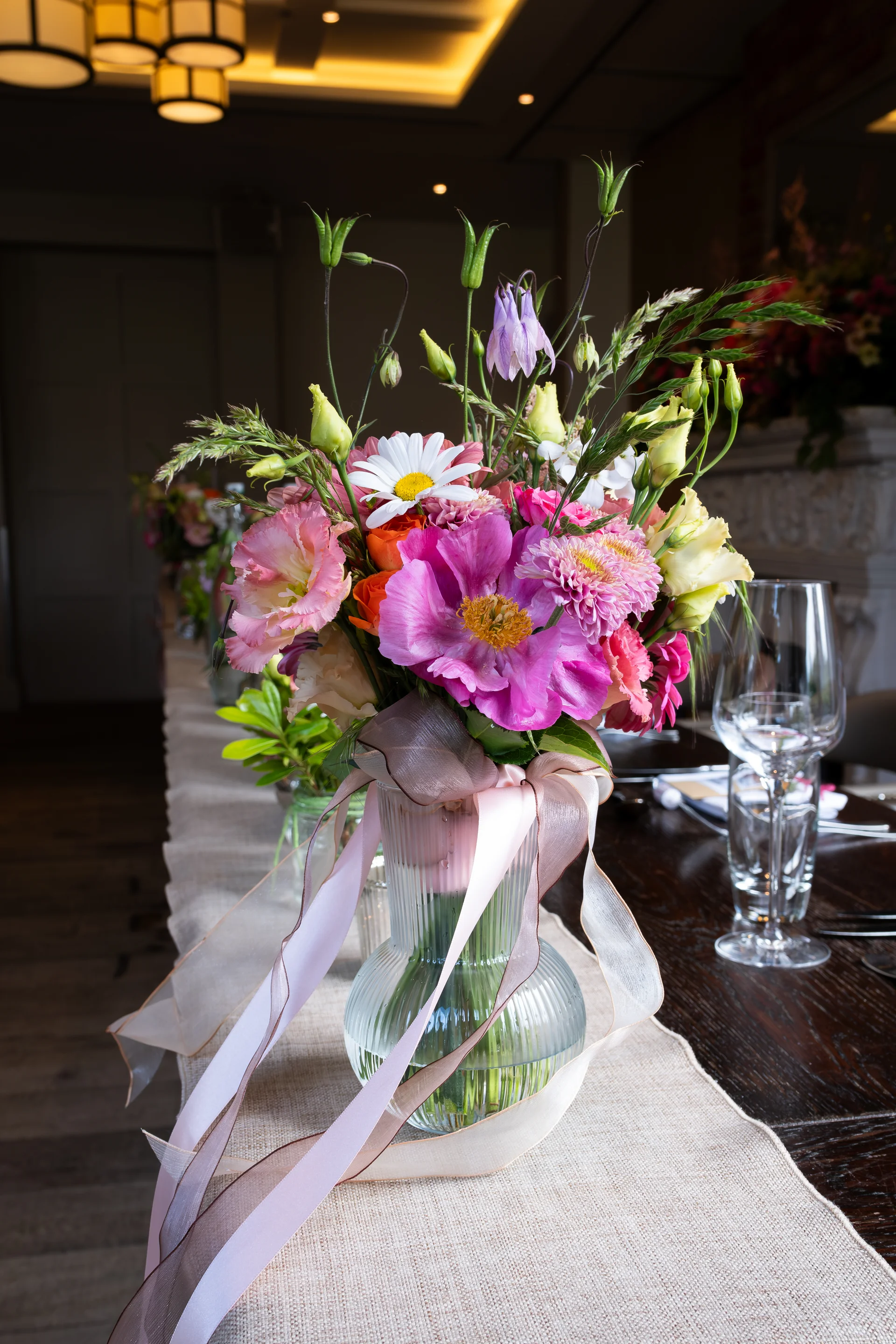 A vibrant garden-style floral centrepiece featuring pink and white blooms in a ribbed glass vase sits atop a long wooden reception table.