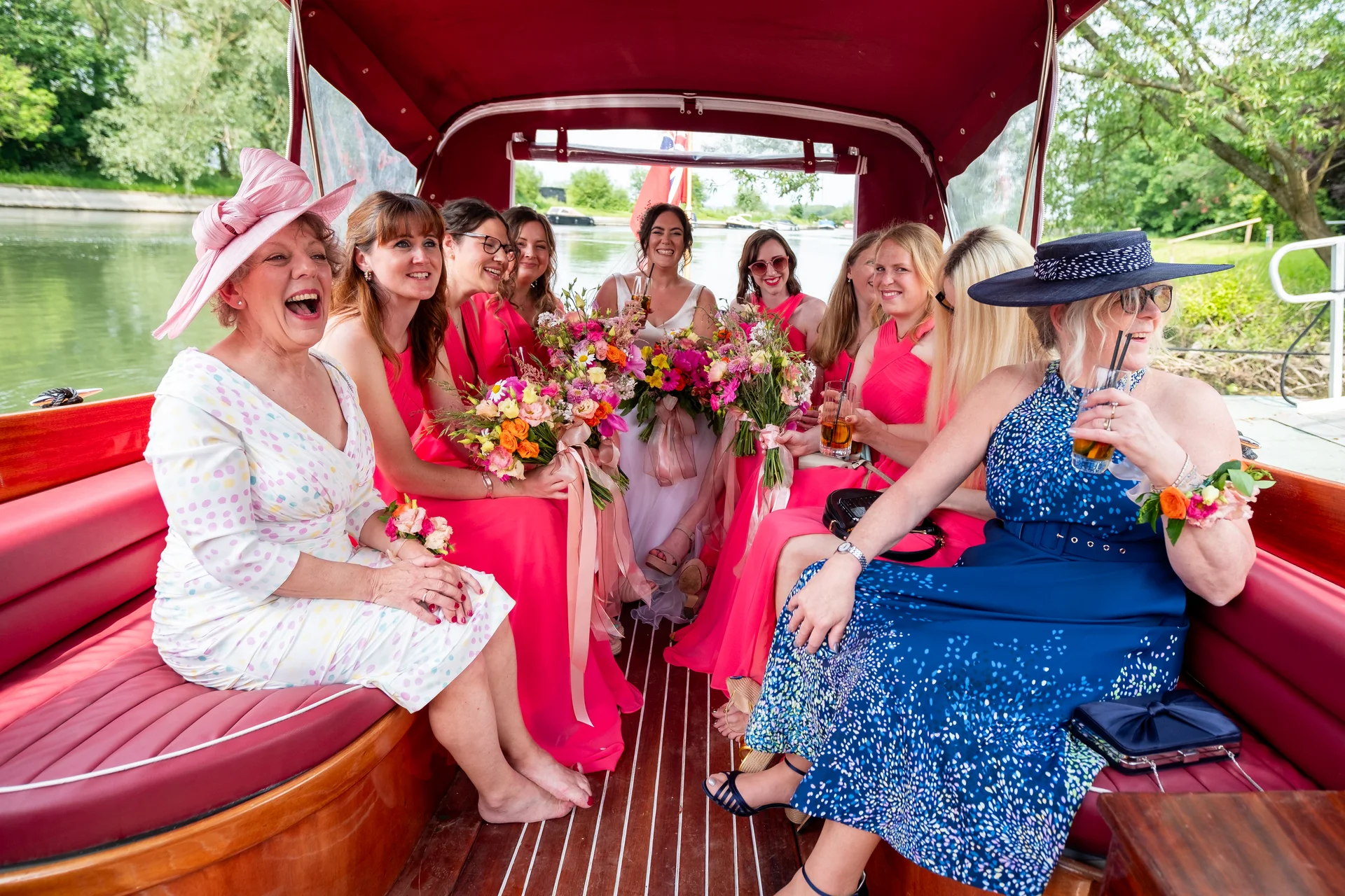 A bride and her bridesmaids celebrate aboard a canopied boat on the water, laughing and holding vibrant bouquets while dressed in bright pink and patterned dresses.