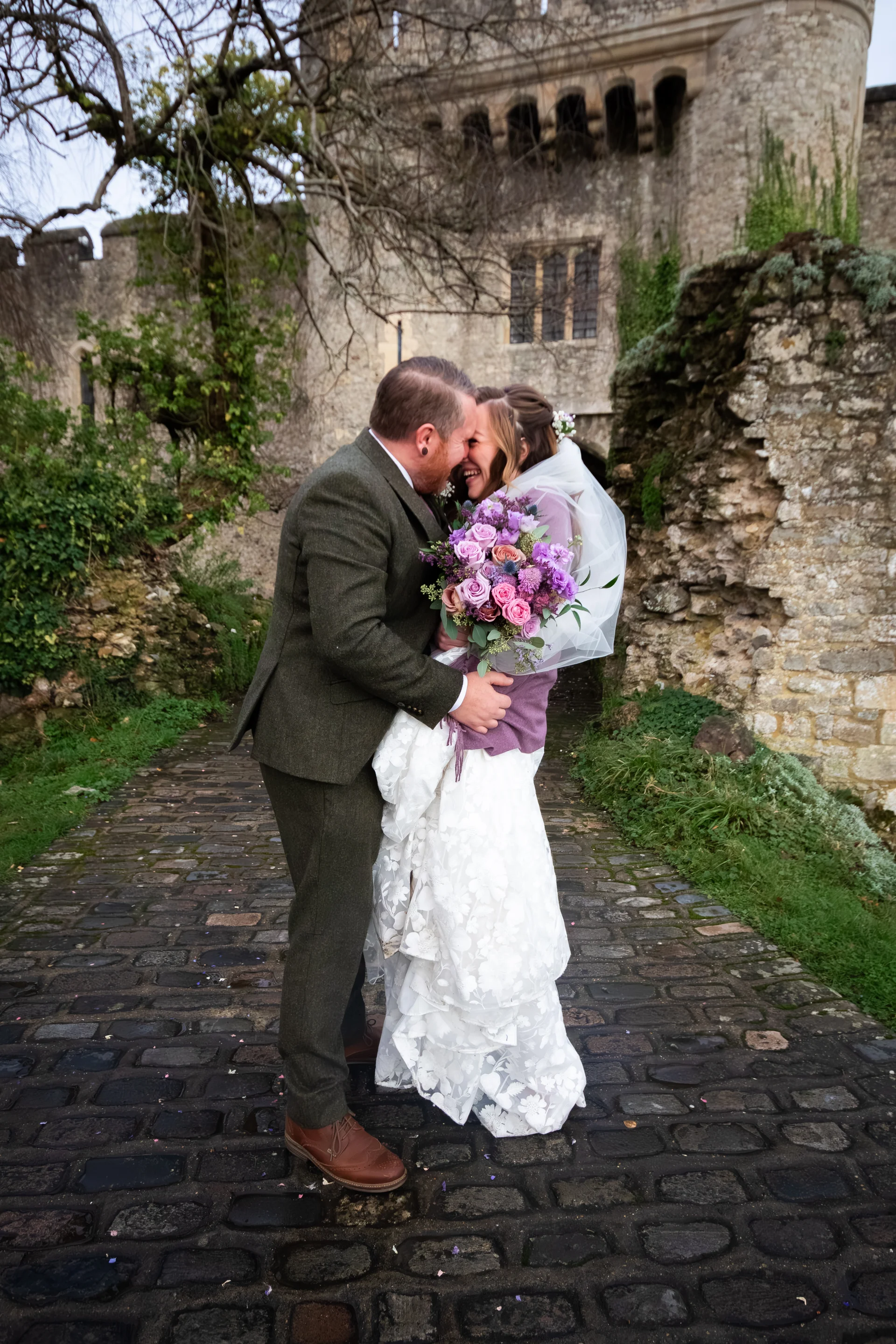 A joyful newlywed couple share a tender forehead kiss on a cobbled path outside an ancient stone castle, with the bride holding a colourful hand-tied bouquet.