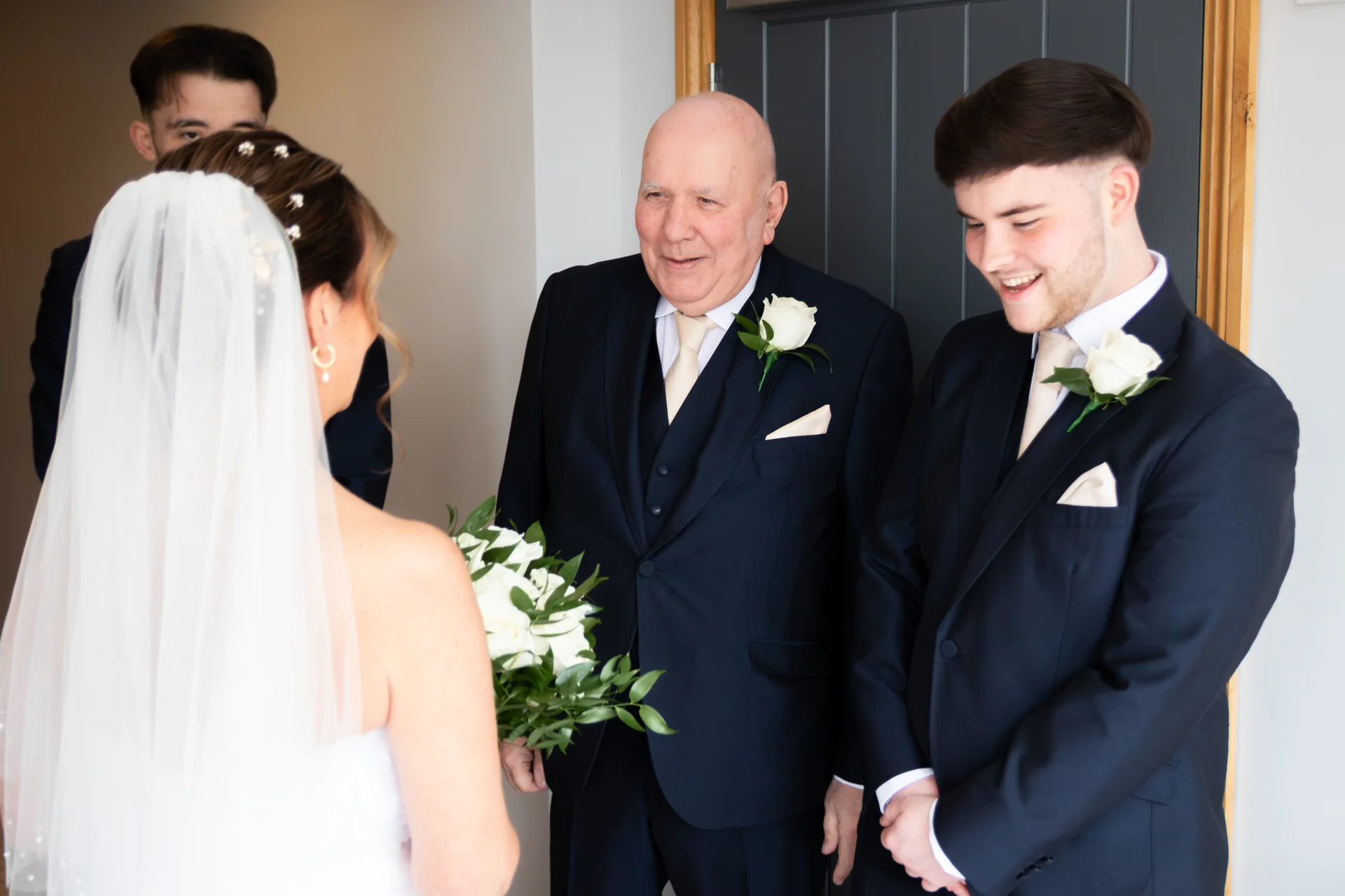 A bride in a white veil and gown holds a bouquet while sharing a warm moment with an older family member and smiling groomsmen in navy suits.