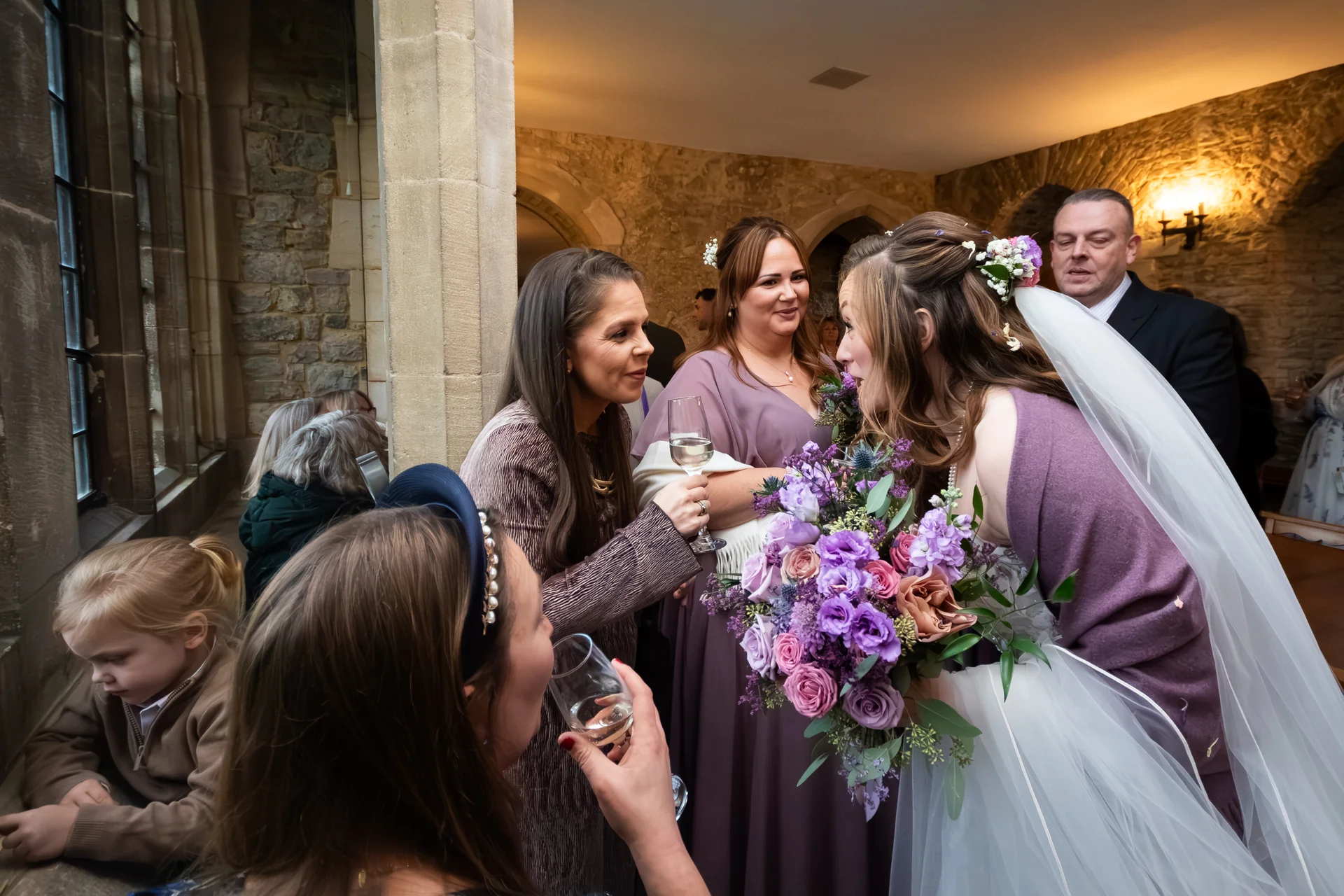 A bride in a veil and purple wrap holds a large purple floral bouquet while chatting with guests holding glasses of wine inside a stone venue.