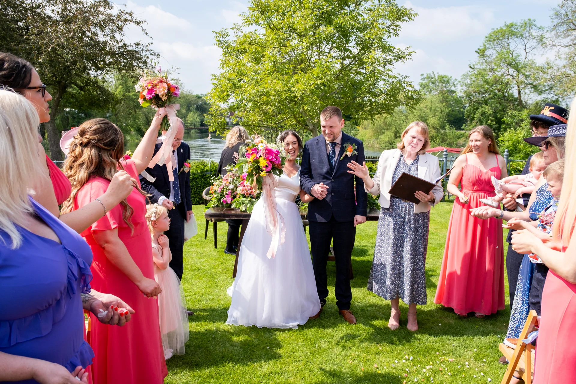 A joyful couple laughs together during an outdoor celebratory ceremony by the water, surrounded by family and bridesmaids in pink dresses.