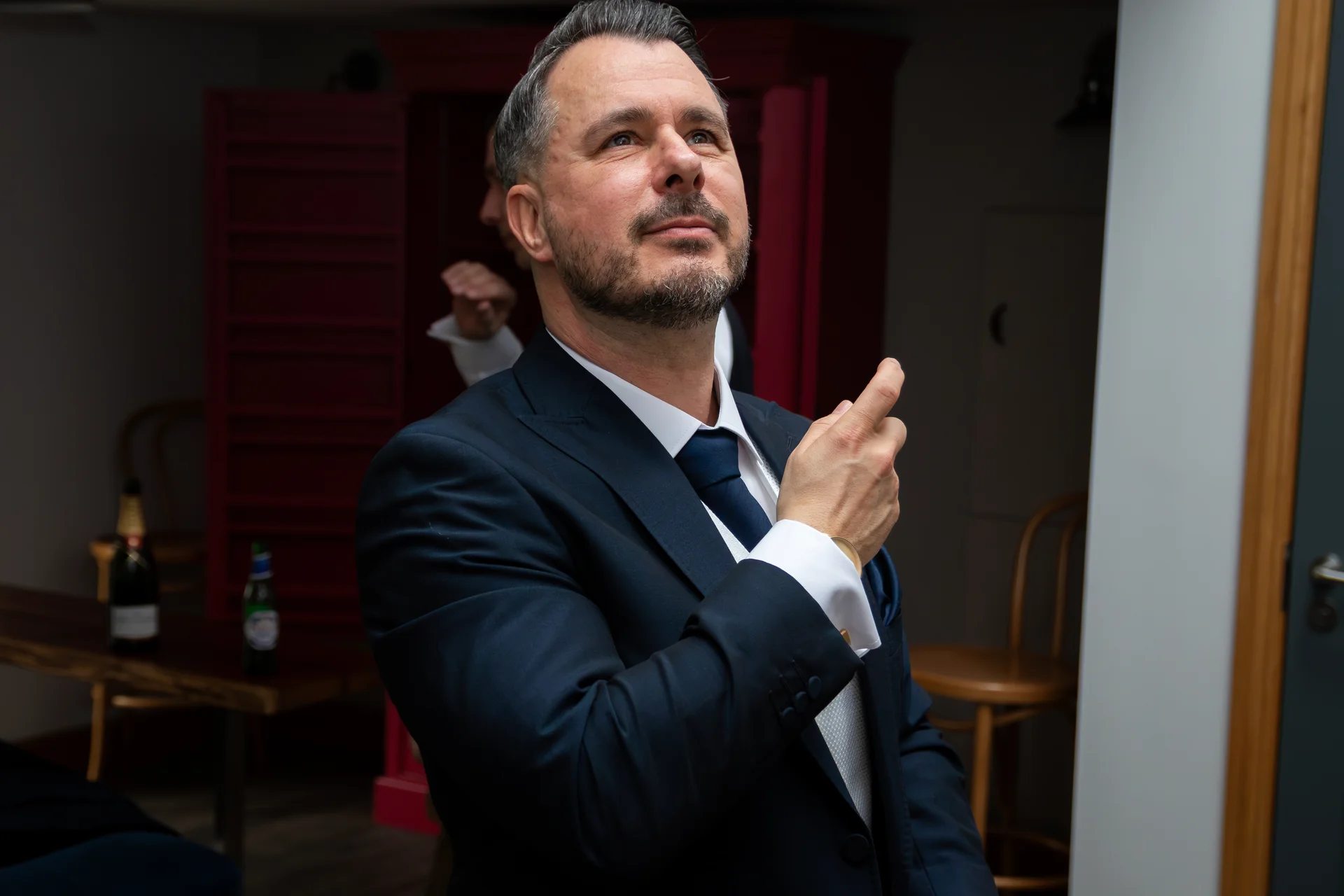 A well-groomed man in a navy suit looks upward thoughtfully while getting ready indoors on his wedding day.