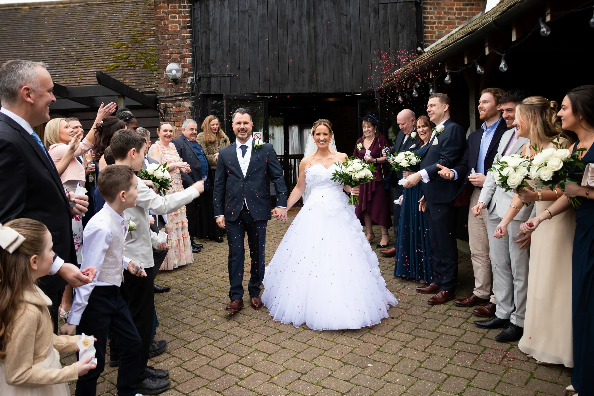 Newlyweds walk hand in hand through a joyful confetti shower outside a rustic stone and timber barn, surrounded by smiling wedding guests and bridal party members.