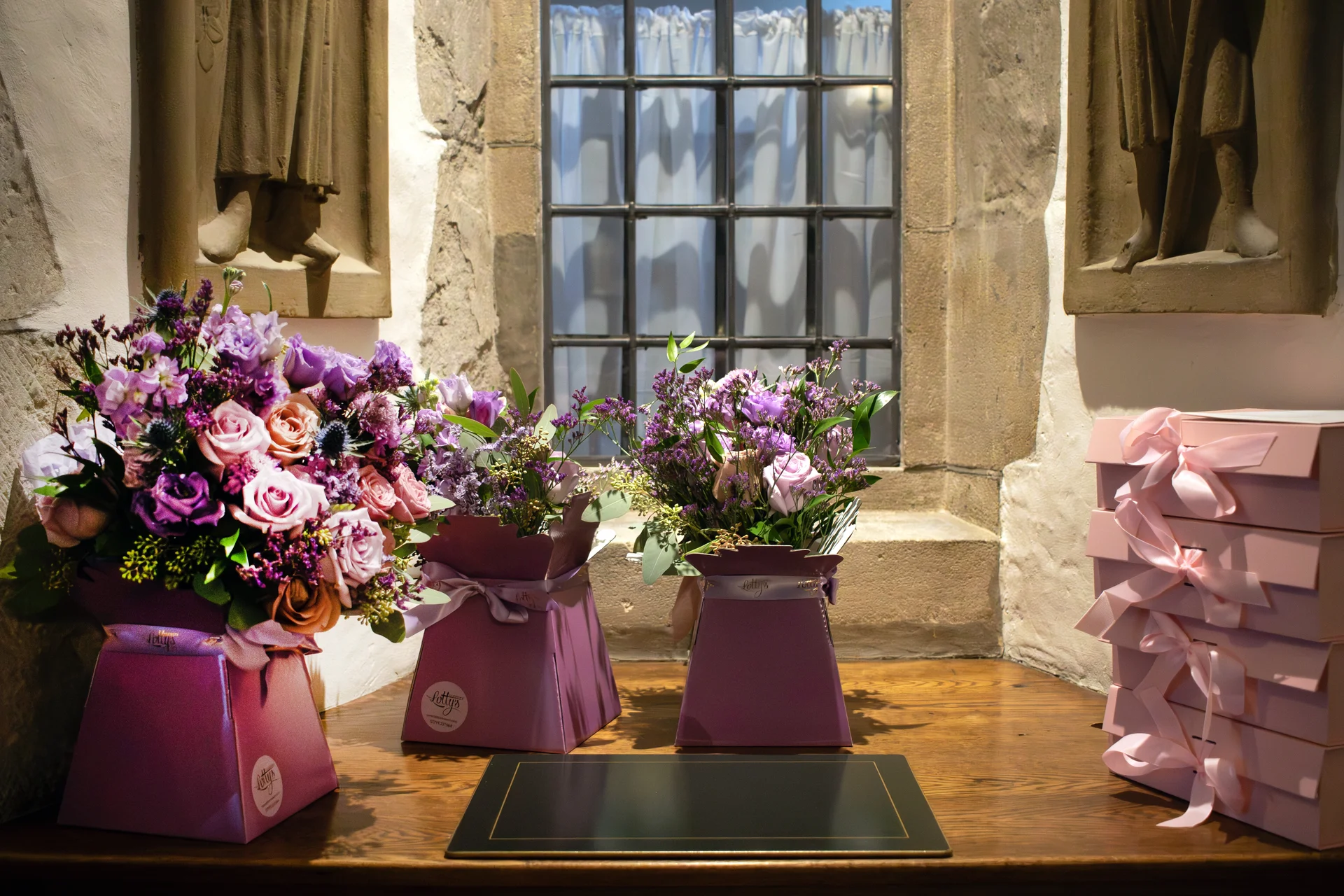 Three purple flower arrangements in matching boxes sit on a wooden table next to a stack of pink gift boxes under a stone window.