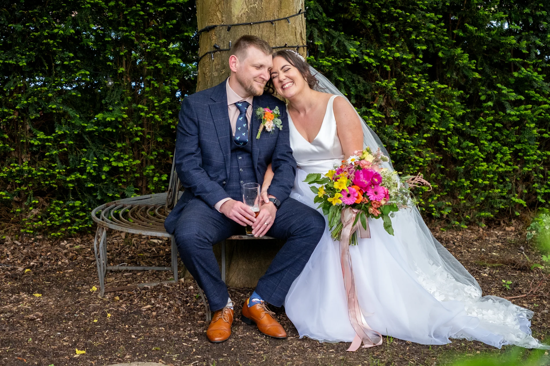 A joyful newlywed couple sits smiling together on a circular bench wrapped around a tree trunk, with the bride holding a colourful floral bouquet.