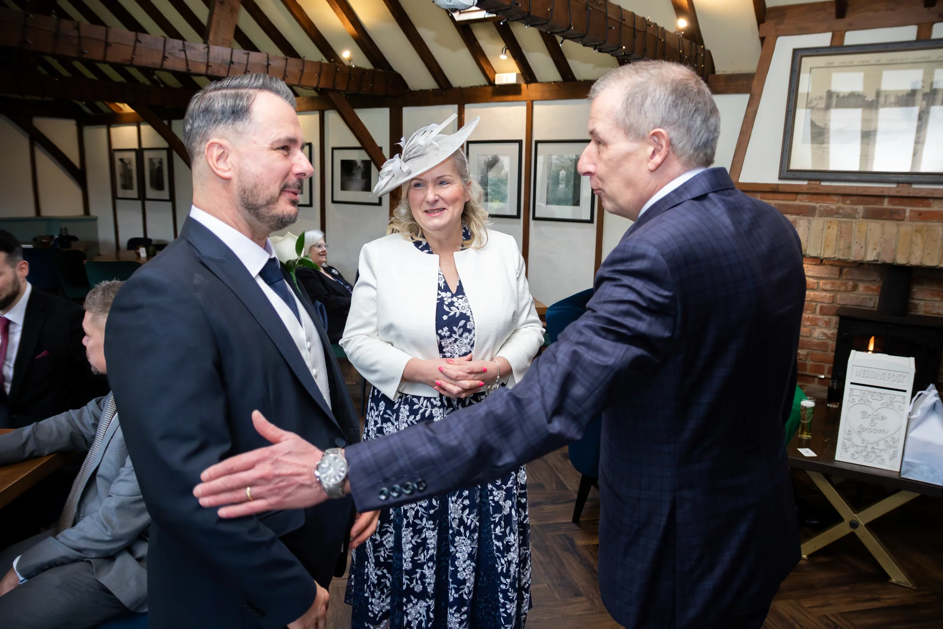 Wedding guests mingle indoors within a rustic barn featuring exposed wooden beams, while a man in a patterned suit welcomes another guest warmly.