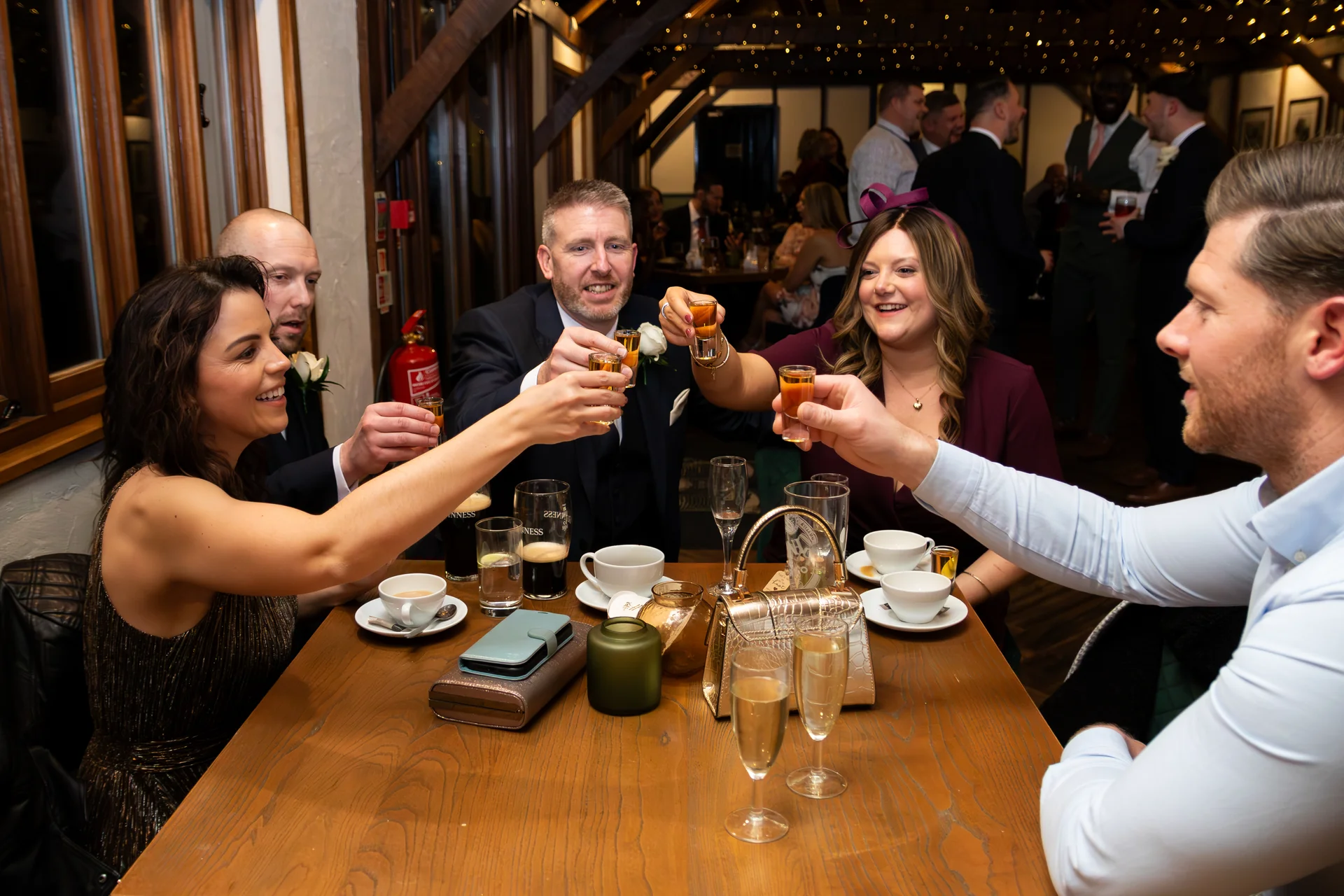 A group of wedding guests cheerfully clink shot glasses together around a wooden table during a lively reception in a rustic pub venue.
