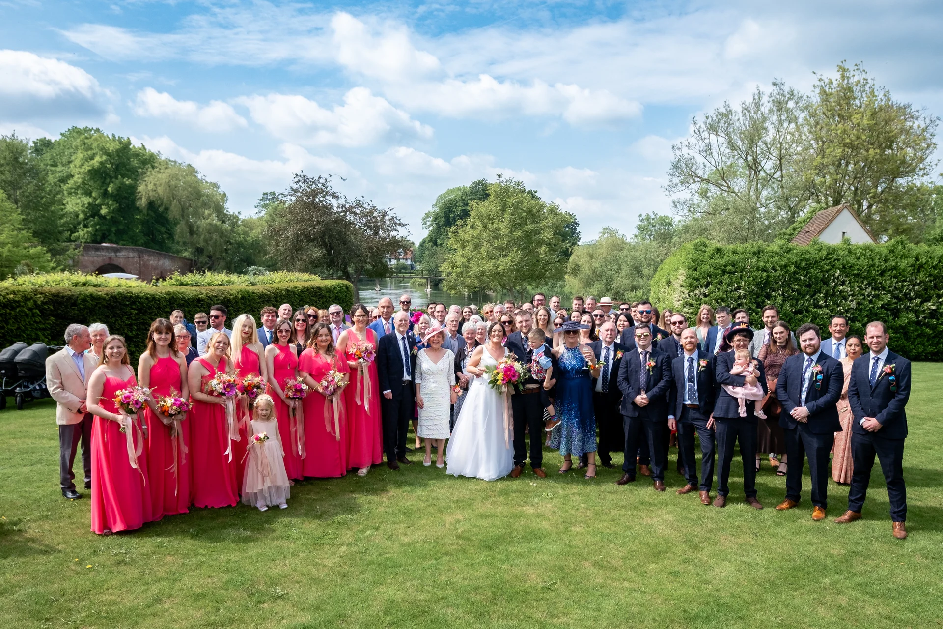 A large wedding party and guests gather outdoors on a manicured lawn by a river under a bright blue sky.