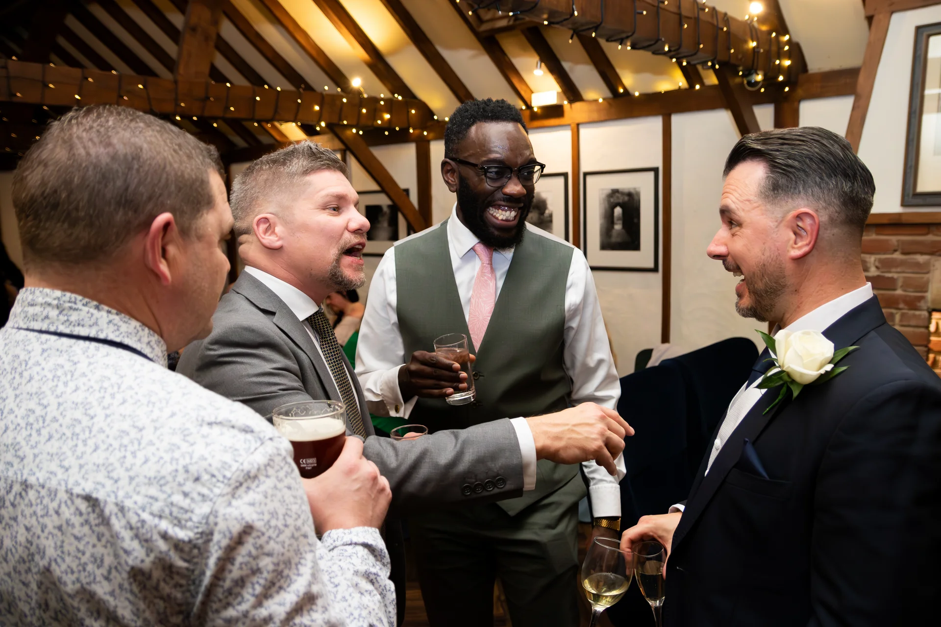 A group of groomsmen and guests laugh and chat animatedly together holding drinks during a wedding reception in a rustic barn venue.