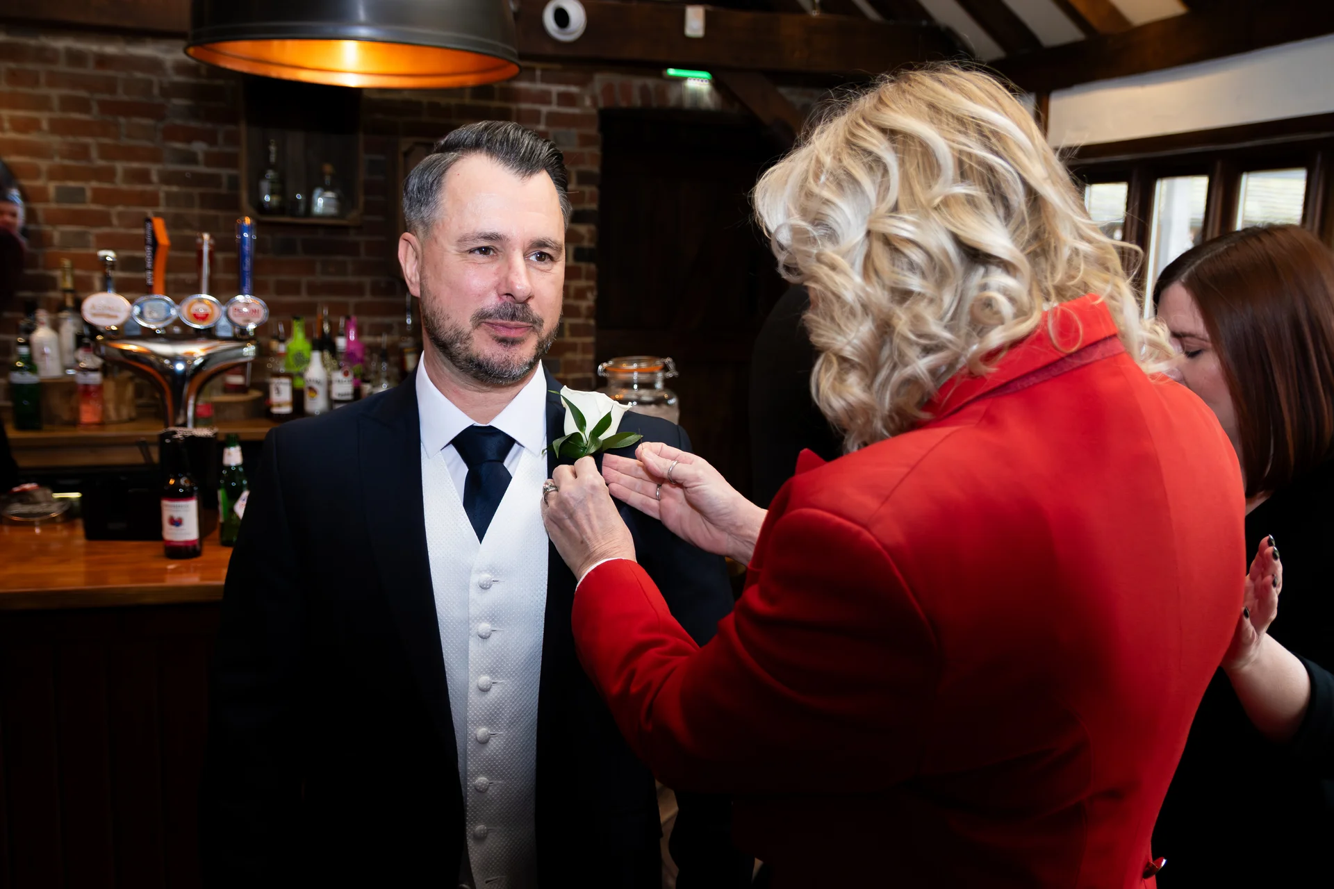 A smiling groom in a black suit and white waistcoat has a buttonhole pinned to his lapel inside a rustic pub setting.