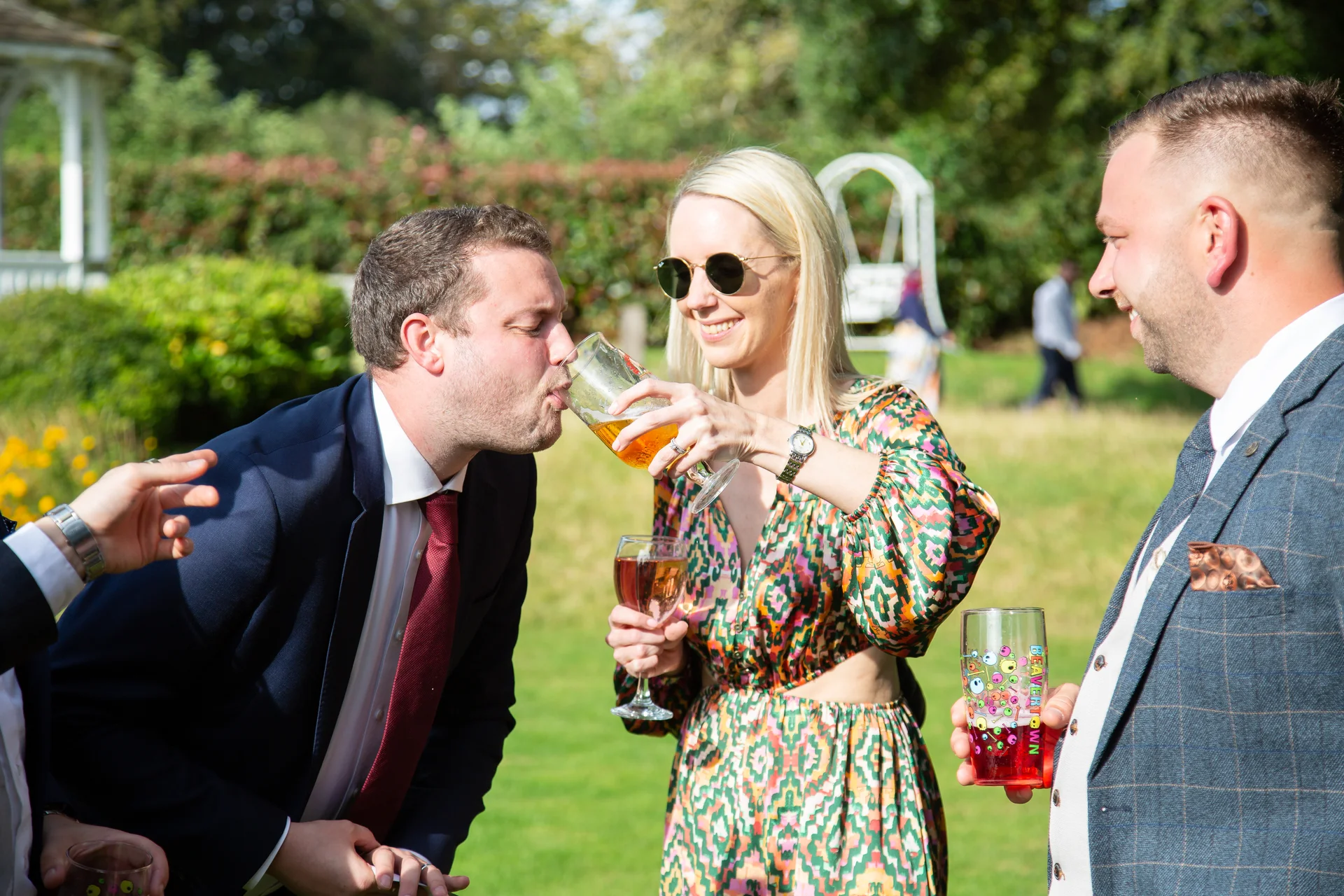 Wedding guests enjoy drinks outdoors during a sunny reception in a garden setting.