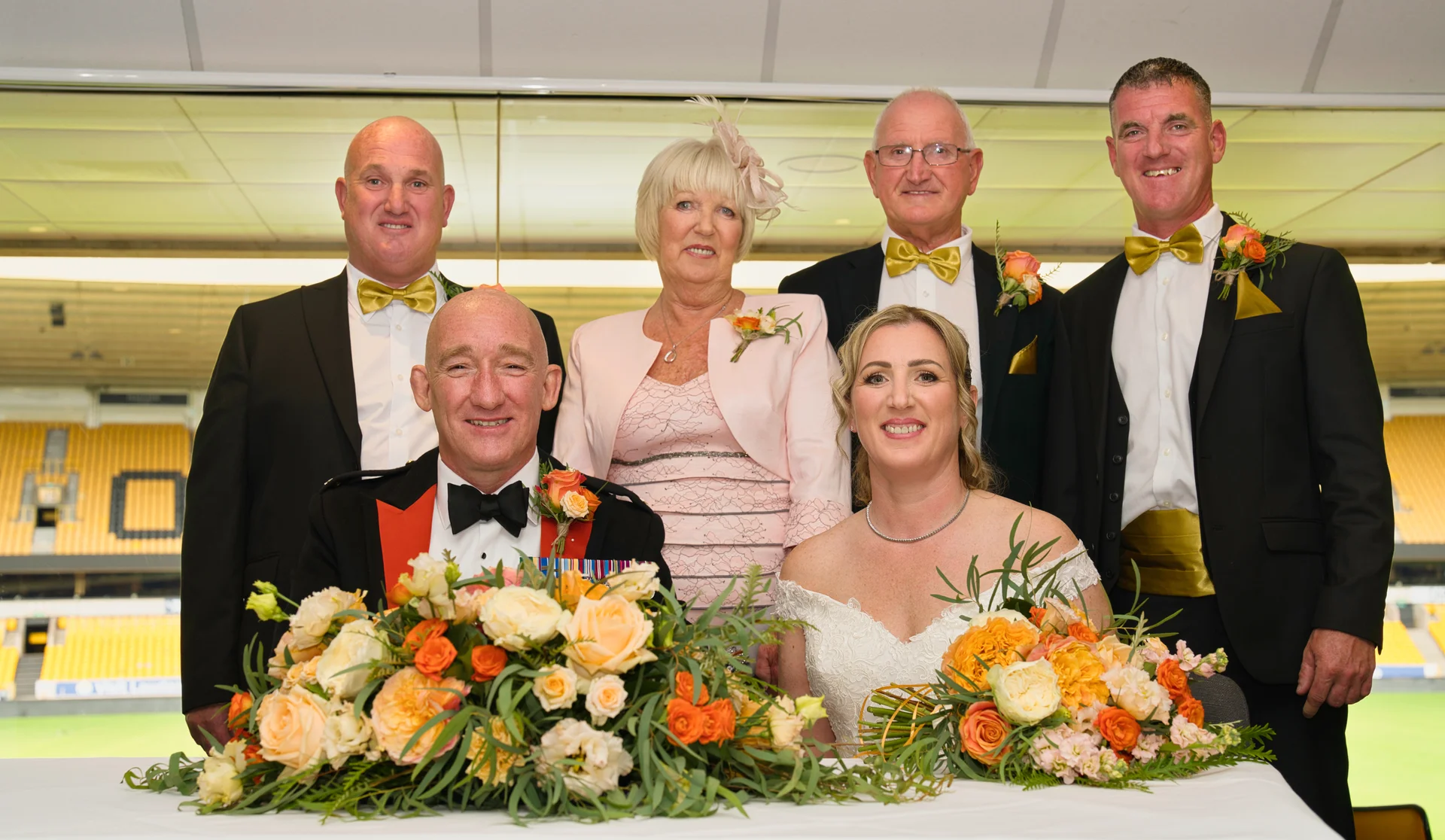 A formal group portrait of the bride, groom in a military uniform, and wedding party seated and standing at the top table in a sports stadium venue.