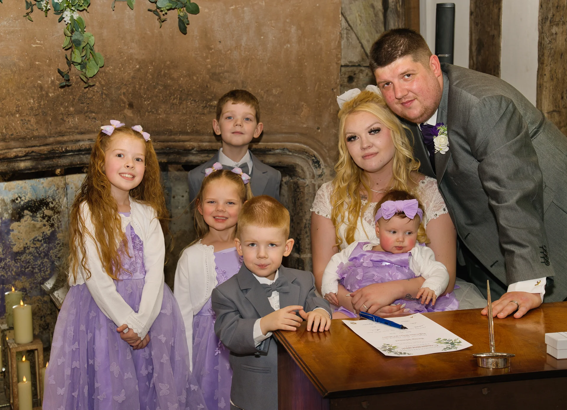 A newly married couple and several children pose together around a wooden desk as the bride and groom sign the register in front of a rustic fireplace.