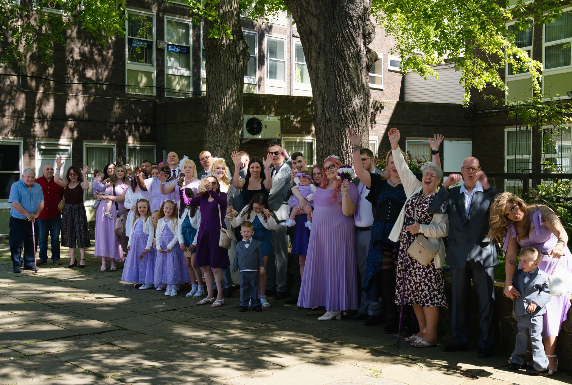 A large wedding group poses and cheers happily together in a sunlit paved courtyard framed by mature trees.