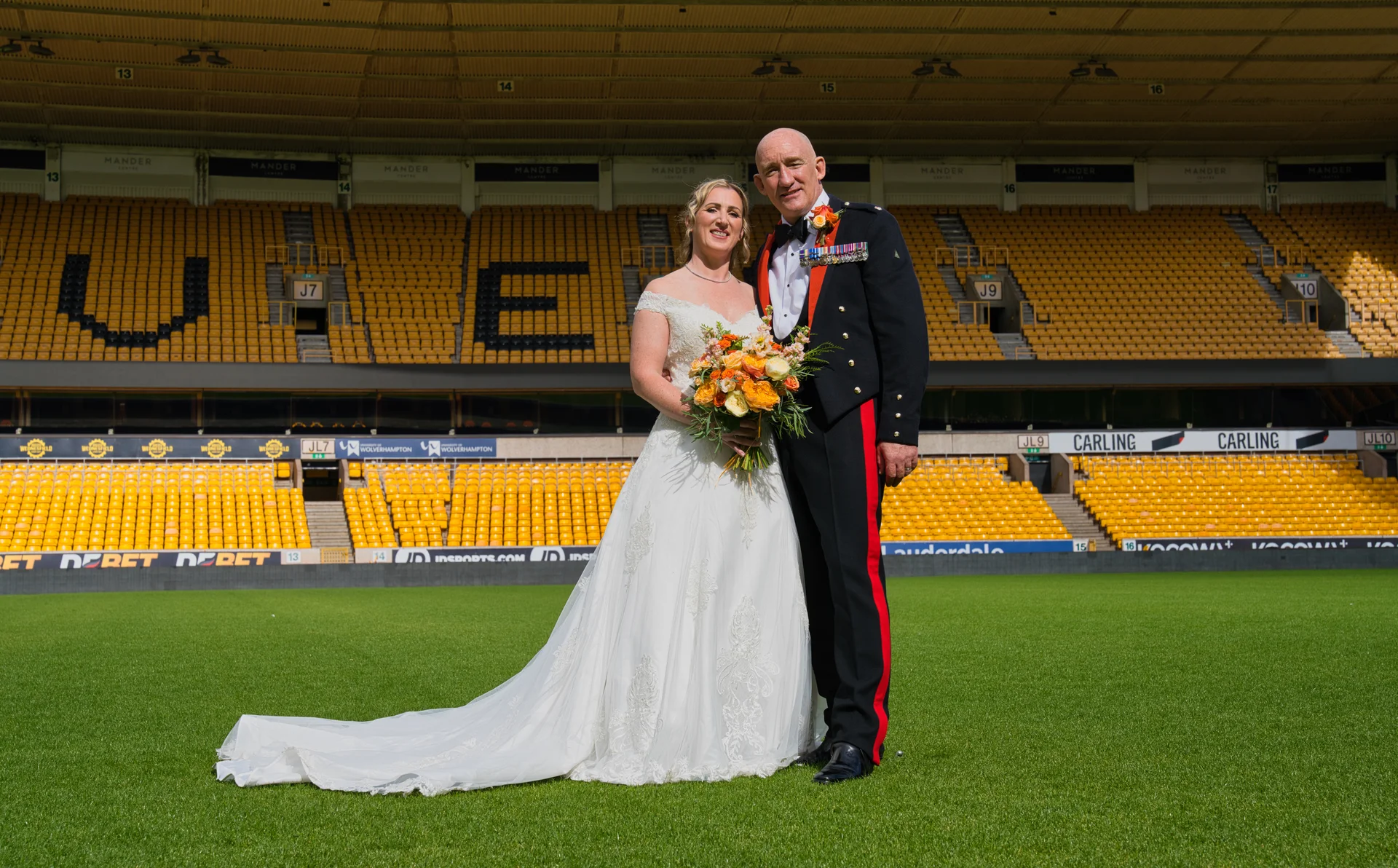 A bride in an a-line lace wedding dress holding a vibrant bouquet stands on the green pitch of a stadium next to a groom wearing a military dress uniform.