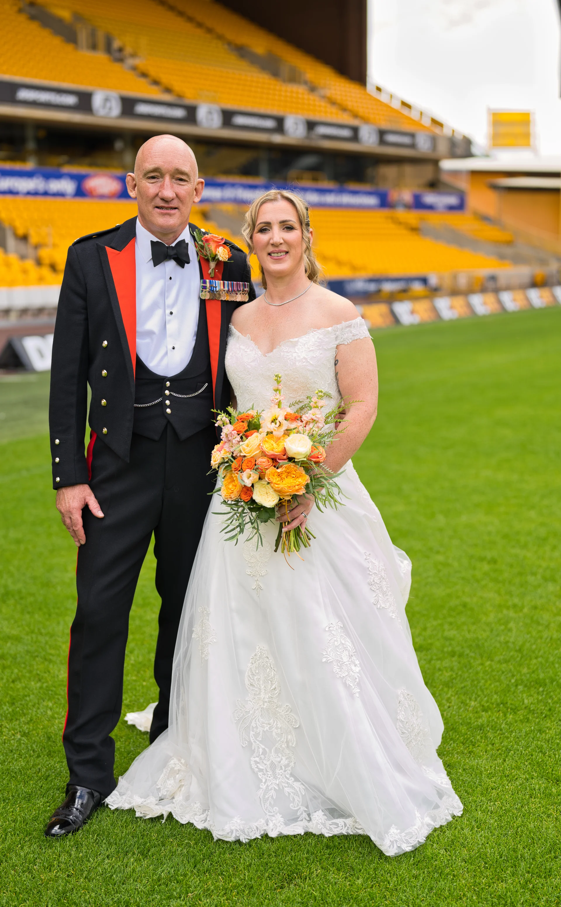 A newlywed couple poses on a vibrant green sports stadium pitch, with the groom in a ceremonial military uniform and medals alongside the bride in an off-the-shoulder lace wedding gown holding a bright orange and yellow floral bouquet.