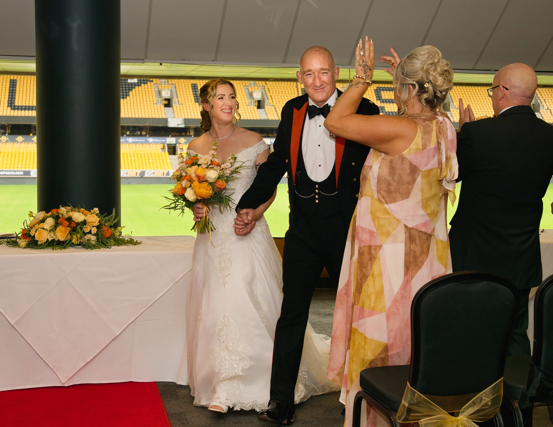 A newlywed couple walk hand-in-hand down the aisle at a sports stadium wedding venue, smiling as cheering guests raise their hands in celebration.