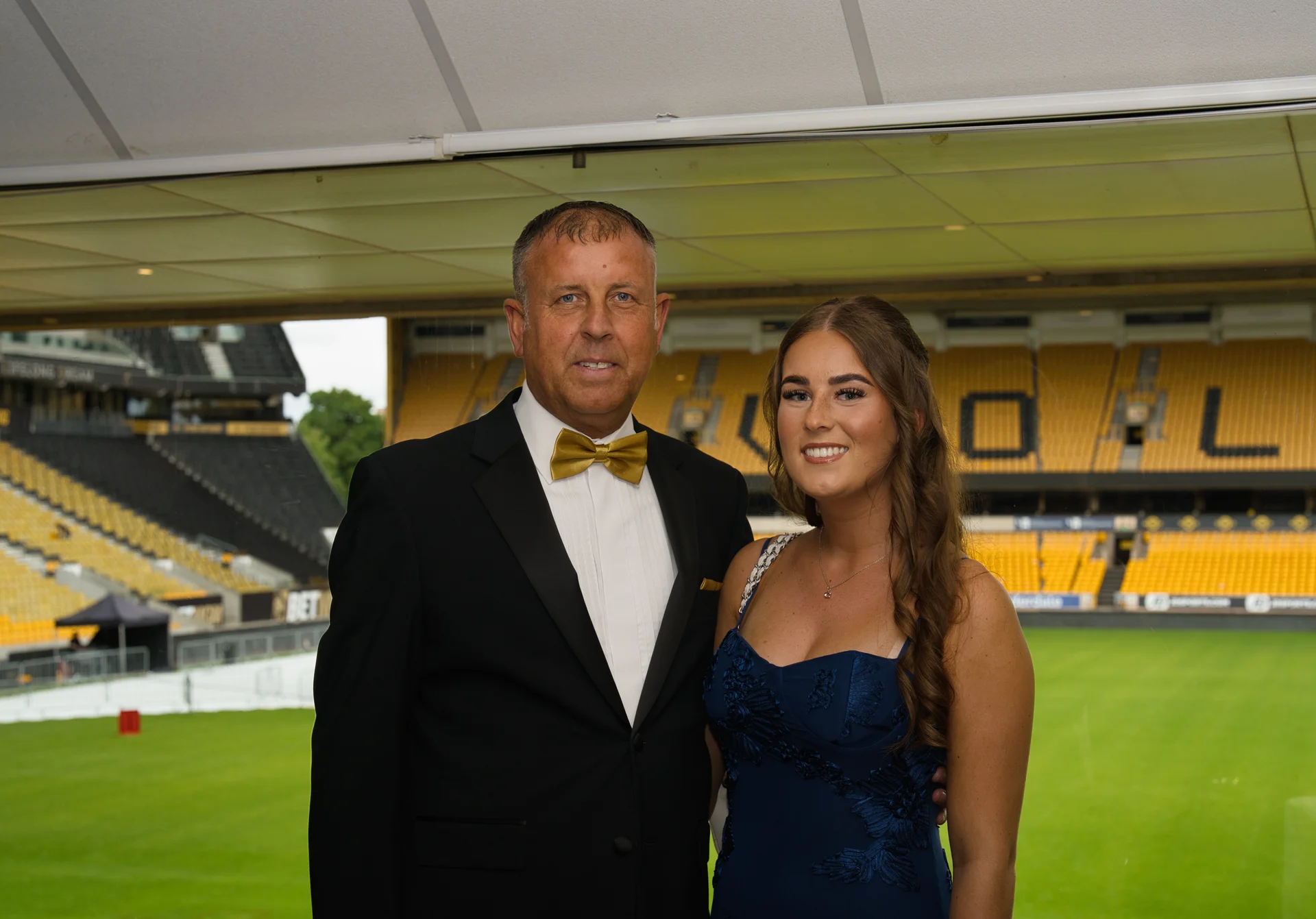 A smiling man in a tuxedo with a gold bow tie and a young woman in a blue dress stand together posing for a portrait inside a football stadium.