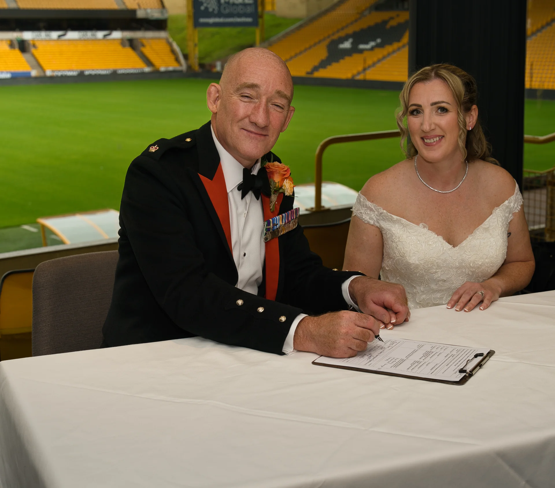 A bride in an off-the-shoulder lace dress and the groom in a military uniform sign the register at a stadium venue.