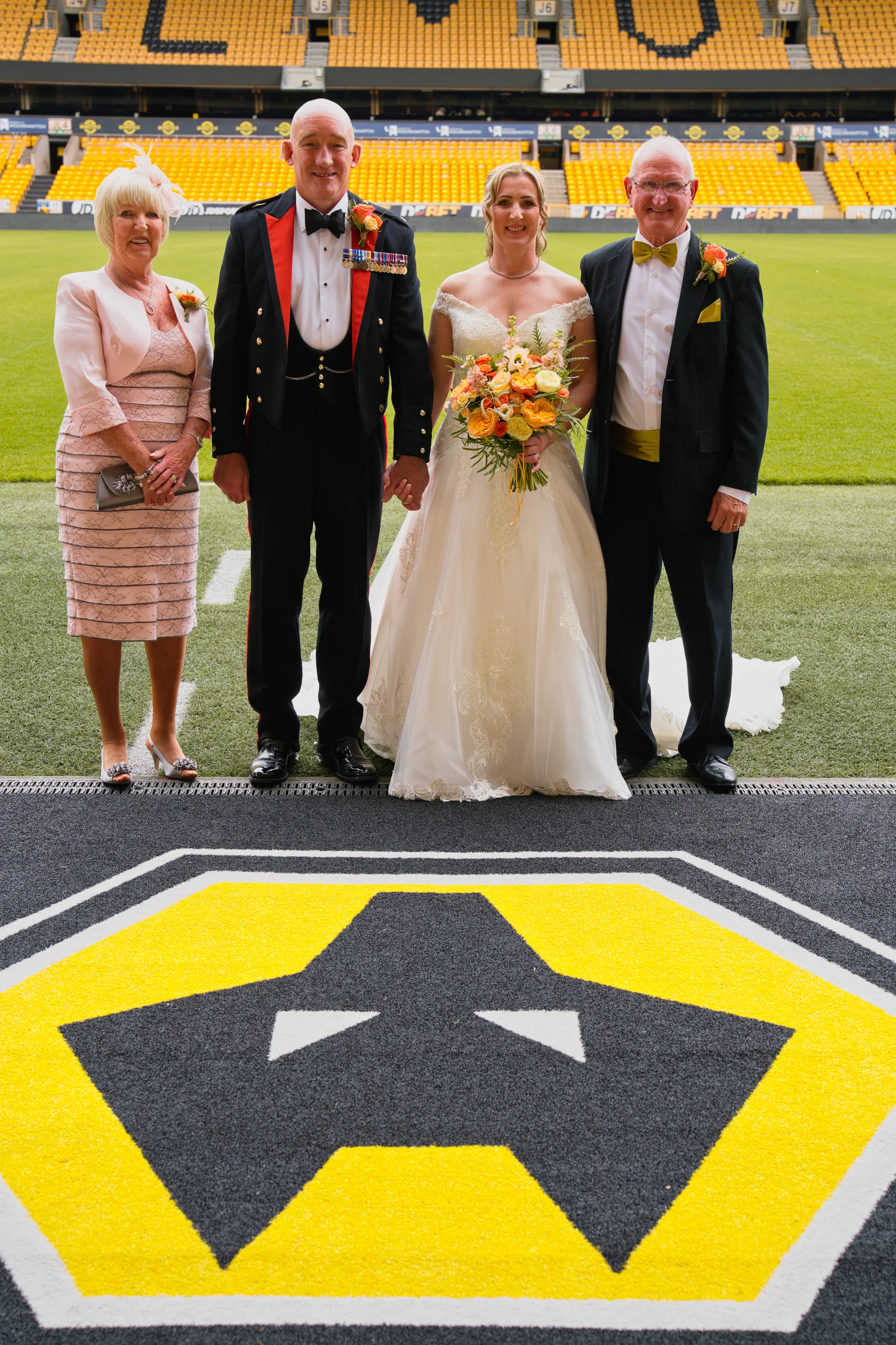 A bridal party poses for a formal group portrait on the edge of a football pitch inside a large sports stadium.