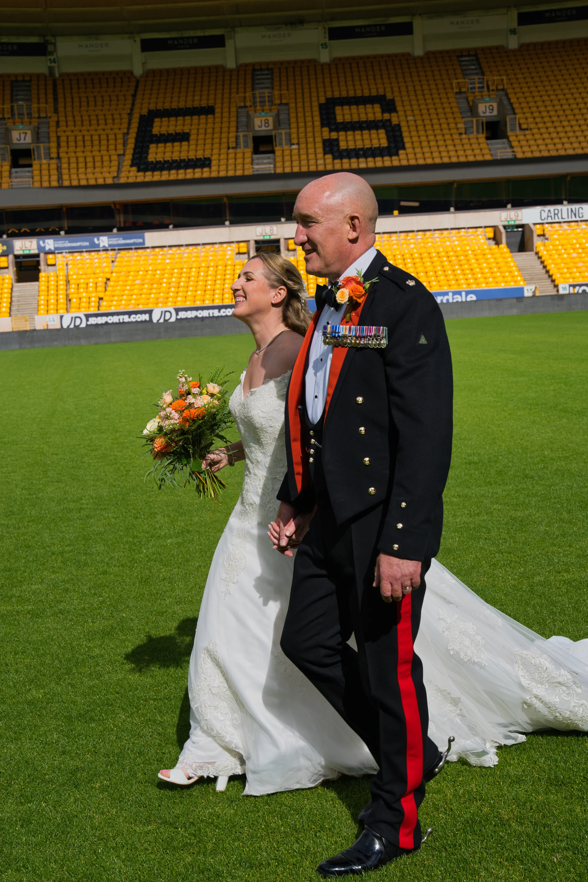 A bride in a lace a-line gown holding an orange bouquet walks hand-in-hand with a groom in a military uniform across the pitch of a sports stadium.