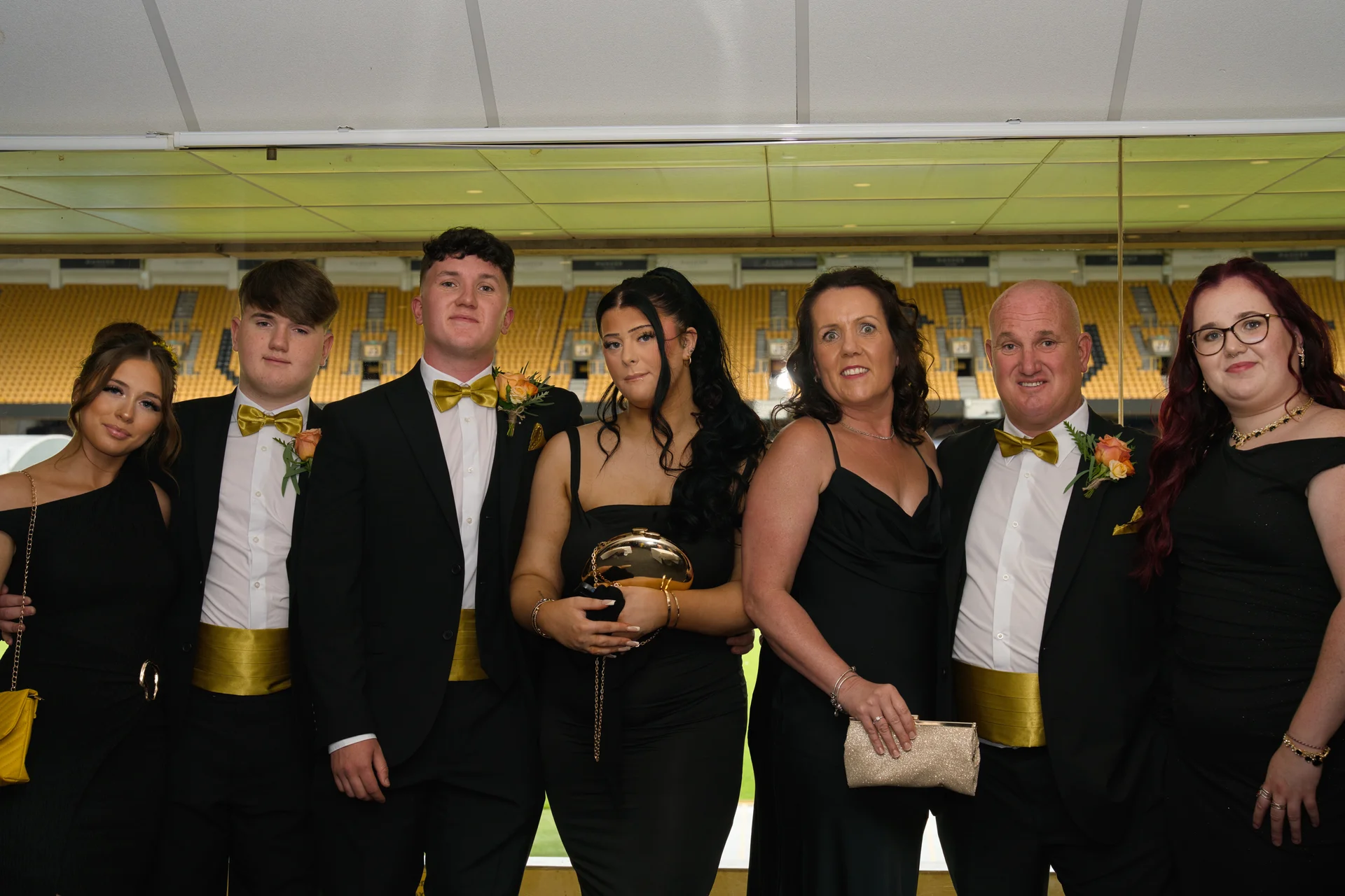 A group of six wedding guests pose together in formal black attire featuring gold cummerbunds and bow ties inside a sports stadium venue.