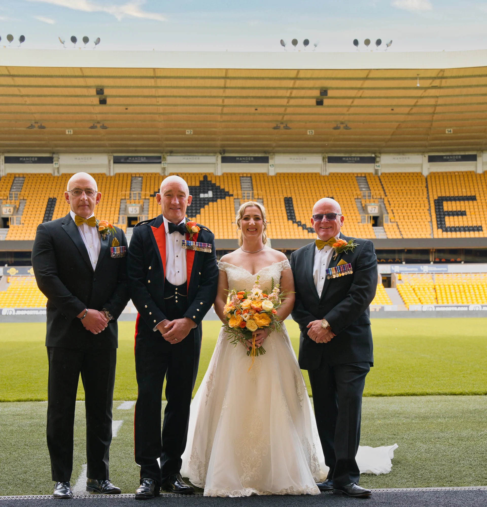 A smiling bride holding a yellow and orange bouquet poses with three men in formal evening wear and military uniforms on the pitch of a large football stadium.