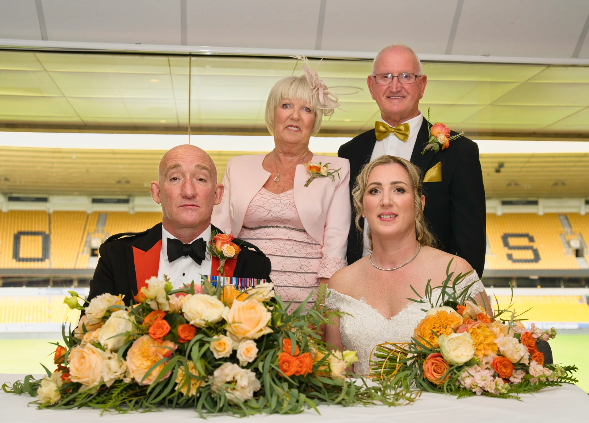 A formal family group portrait featuring a bride in an off-the-shoulder gown, a groom in a military-style mess jacket, and older family members positioned behind a table adorned with vibrant orange and cream floral arrangements inside a sports stadium venue.