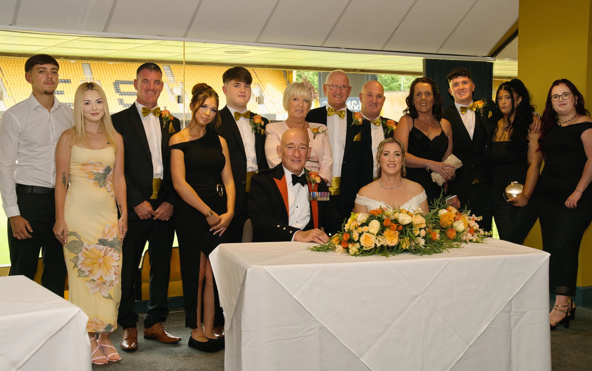 A formal group photo of family and wedding party members posing around the bride and groom seated at a white covered table.