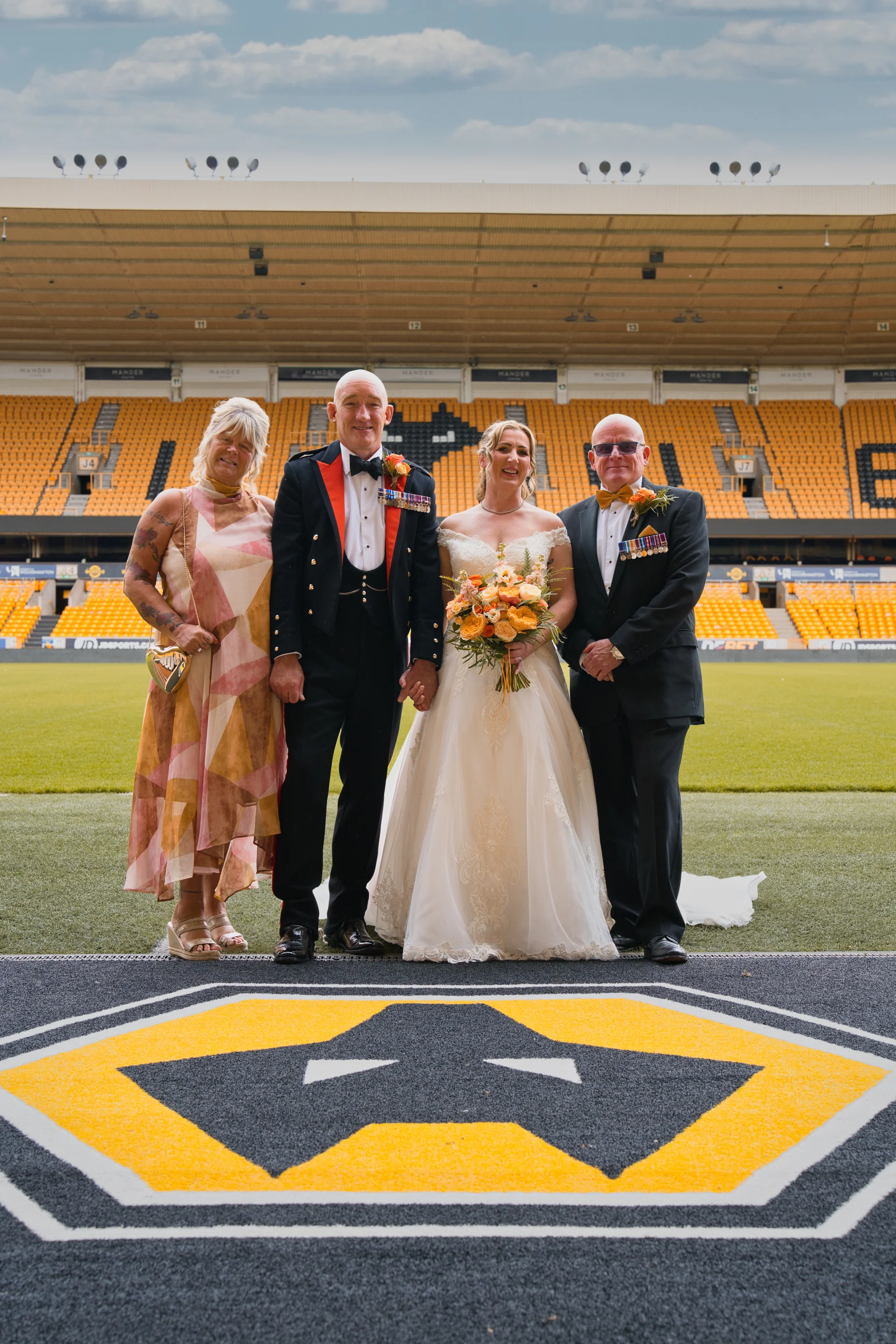 A bridal party poses for a group portrait on the pitch inside a large football stadium with yellow seating in the background.