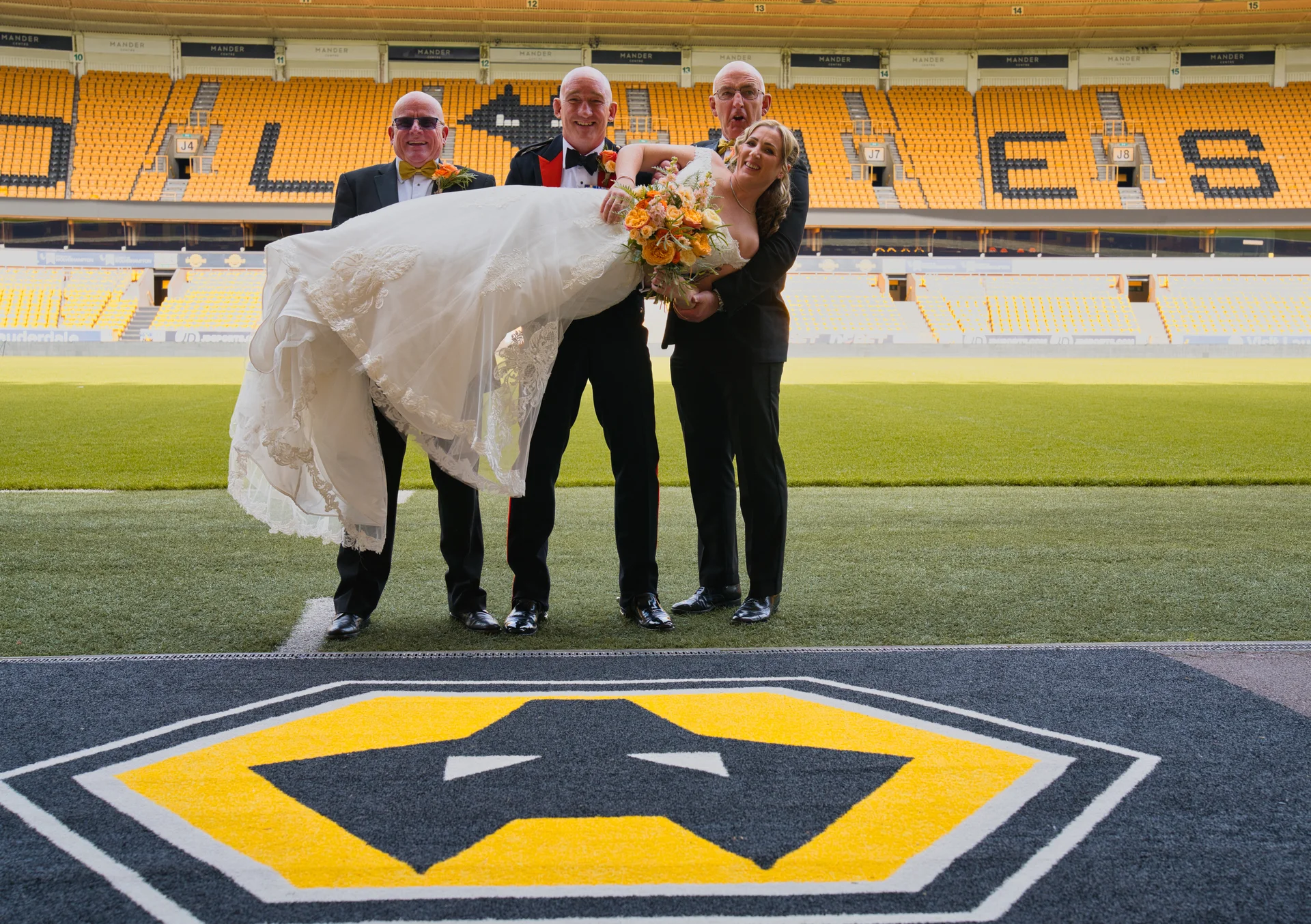 A smiling bride holding her bouquet is lifted horizontally in the air by three men wearing formal suits on the pitch of a large football stadium.