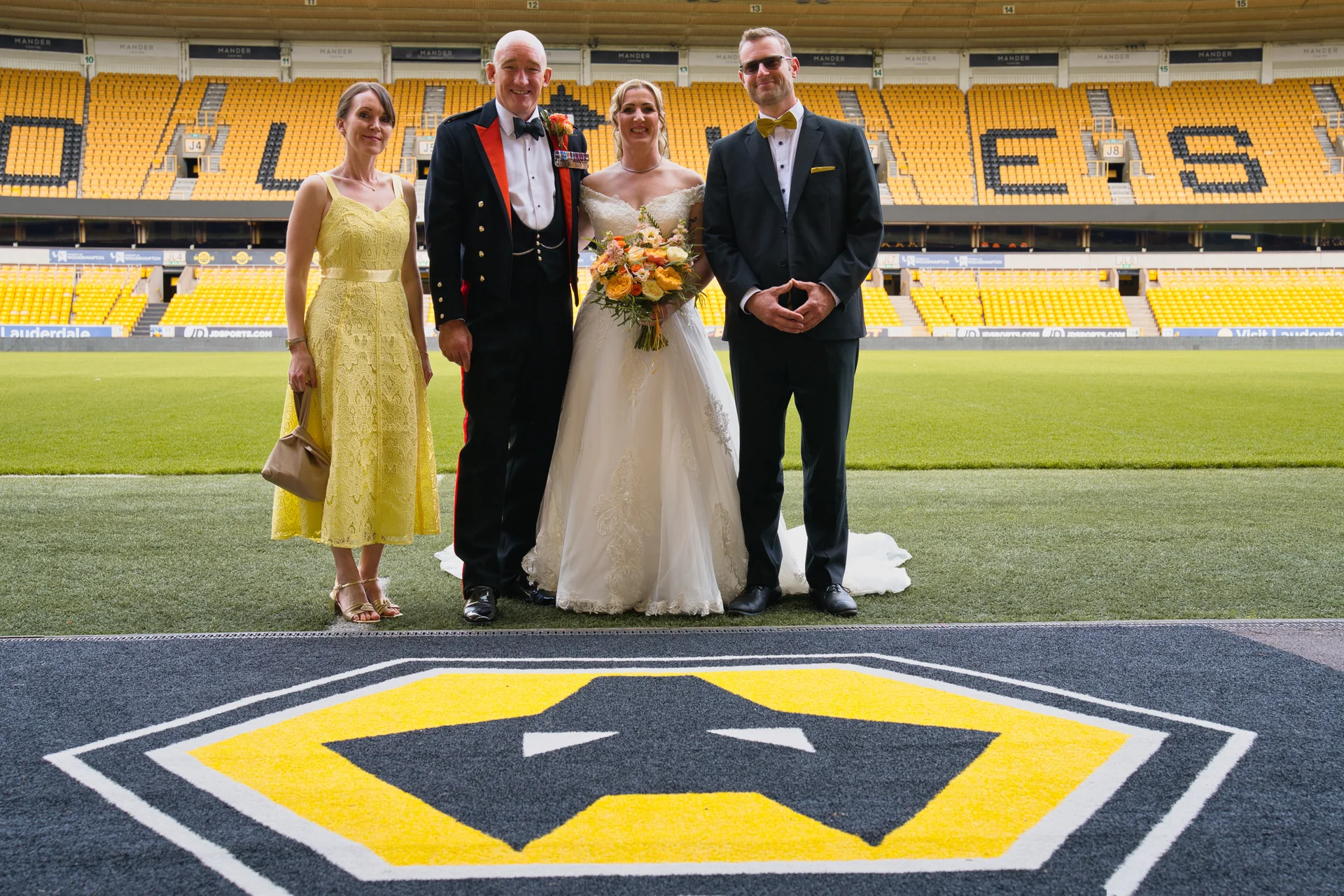 A bridal party poses for a formal group portrait on the pitch inside a large yellow and black sports stadium.