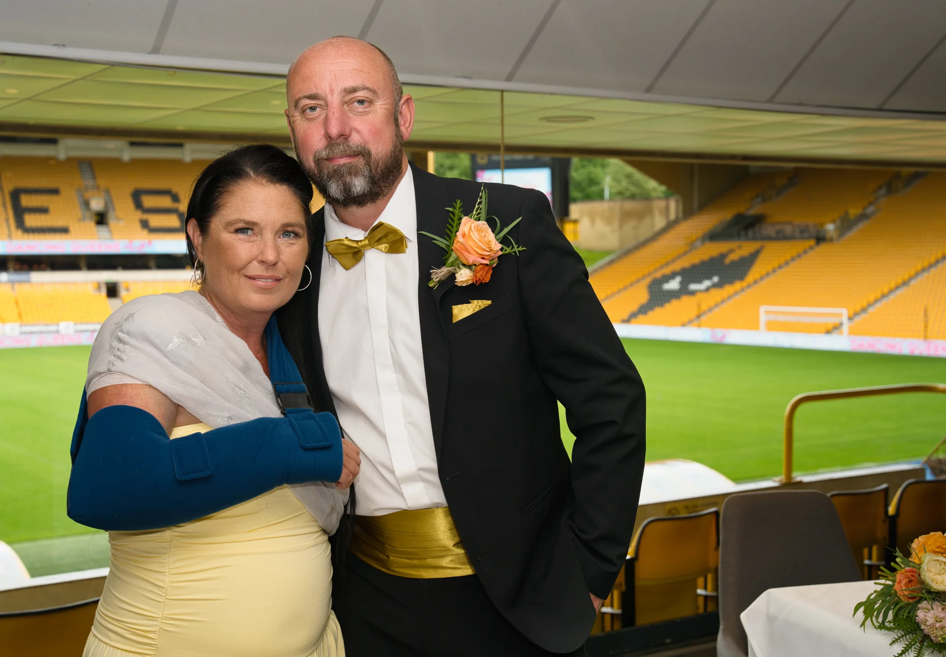 A mature couple poses together inside a sports stadium wedding venue, with the groom wearing a black tuxedo with gold accents and a boutonniere, and the bride wearing a yellow dress and a blue arm sling.
