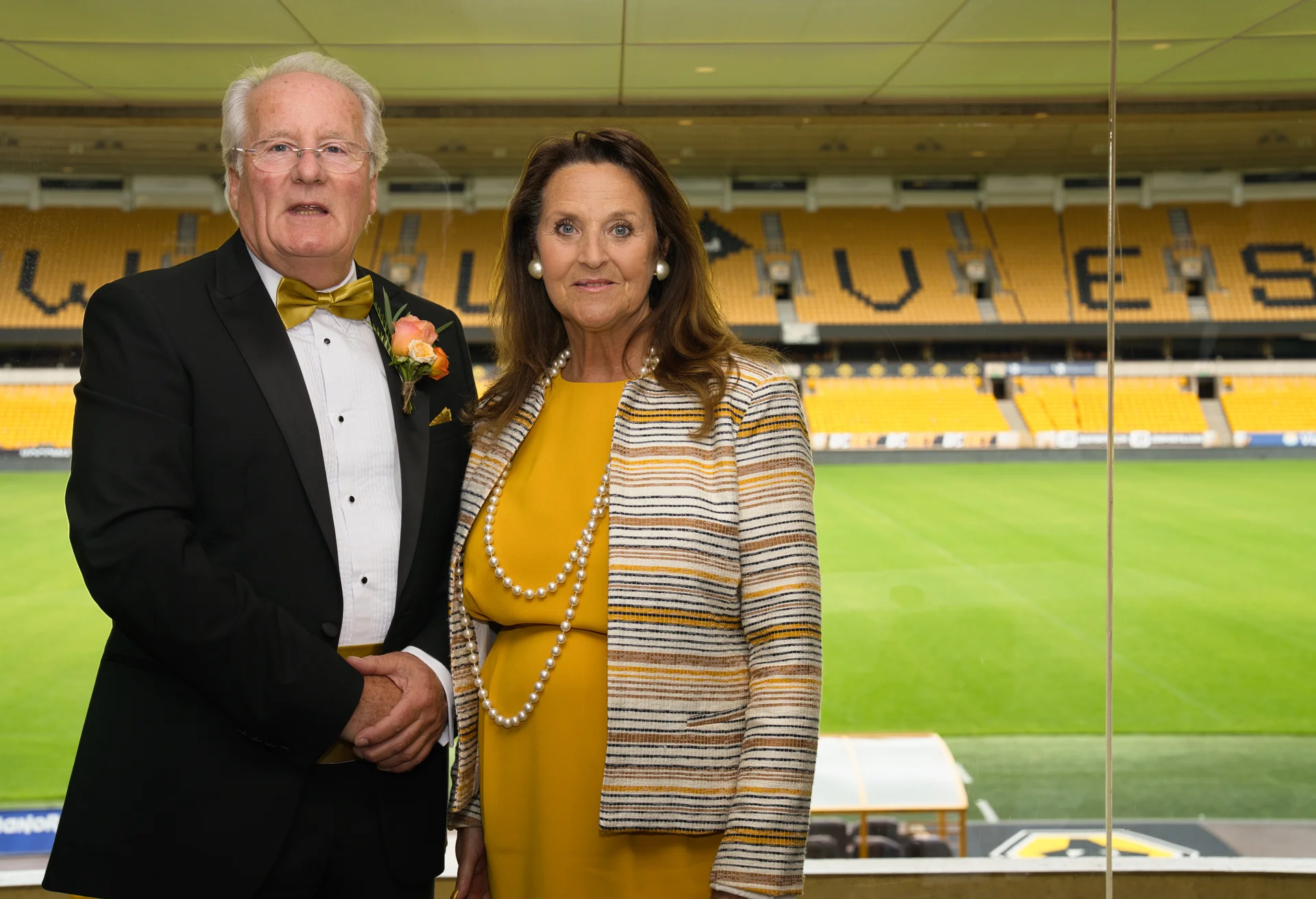 An older couple poses for a portrait inside a luxury suite overlooking a sports stadium pitch.