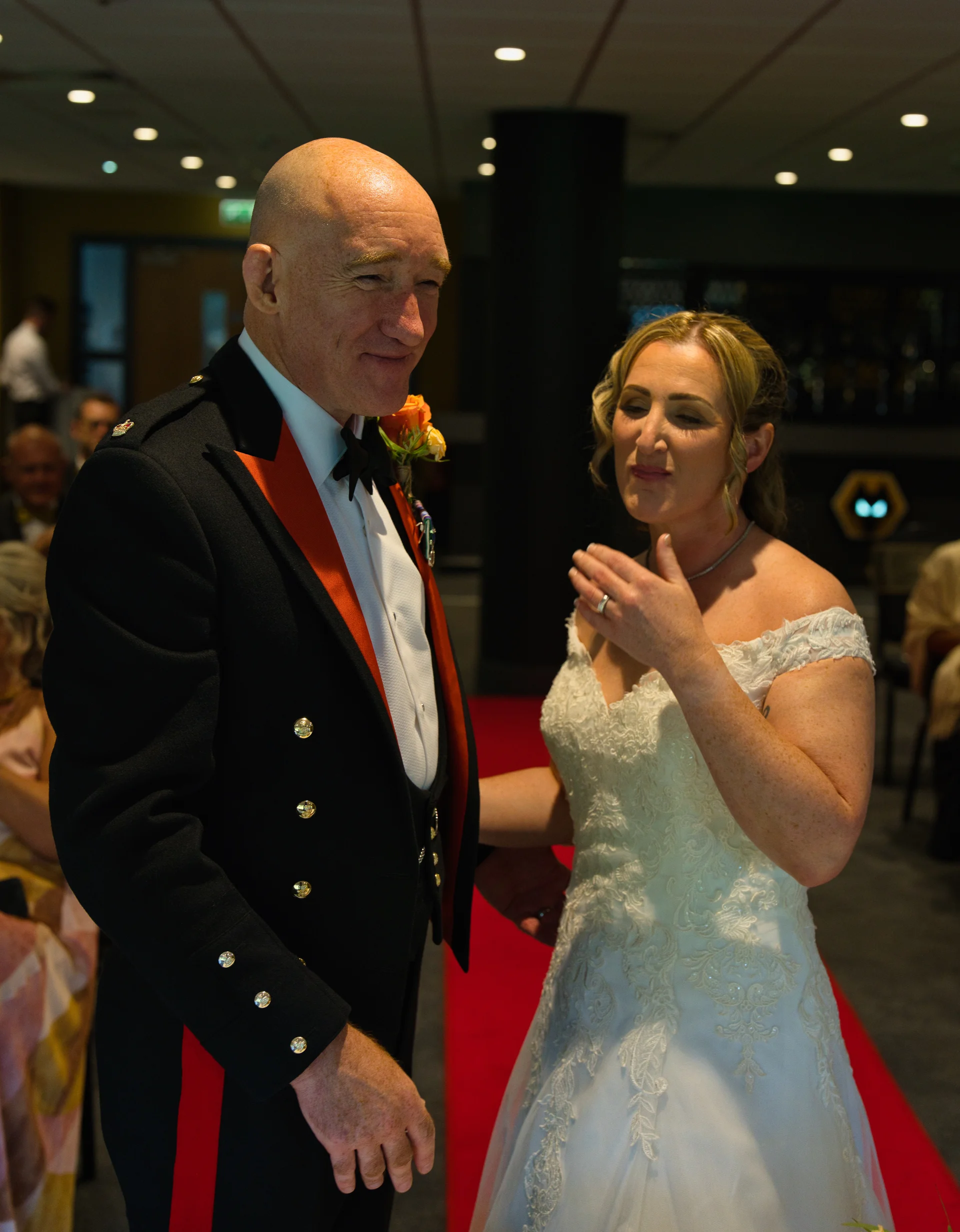 An older groom wearing a military mess dress uniform smiles warmly at his bride as she walks down the aisle in a lace off-the-shoulder wedding gown.