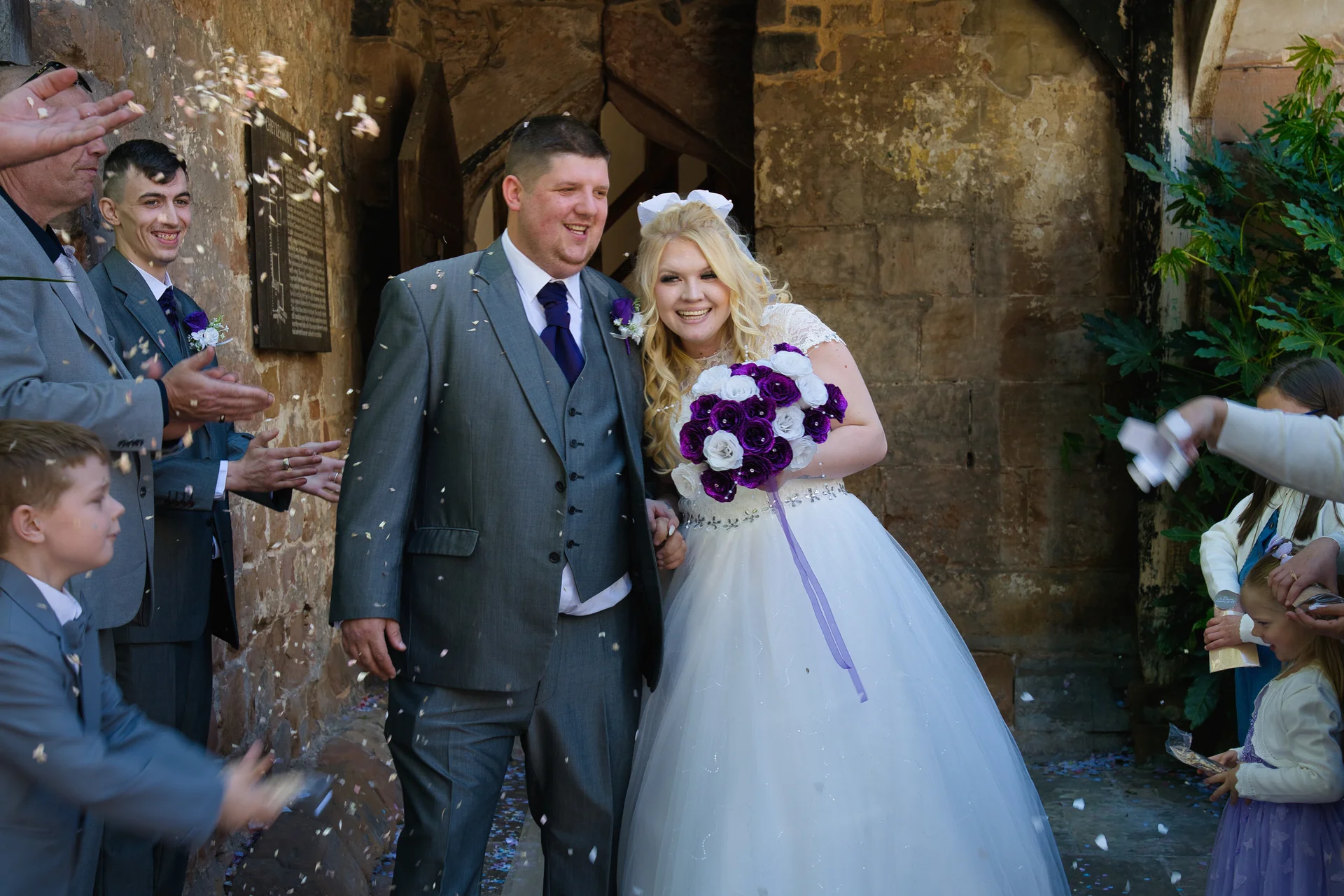 A newlywed couple laugh and smile as they emerge from a historic stone church doorway amidst a shower of confetti thrown by guests.