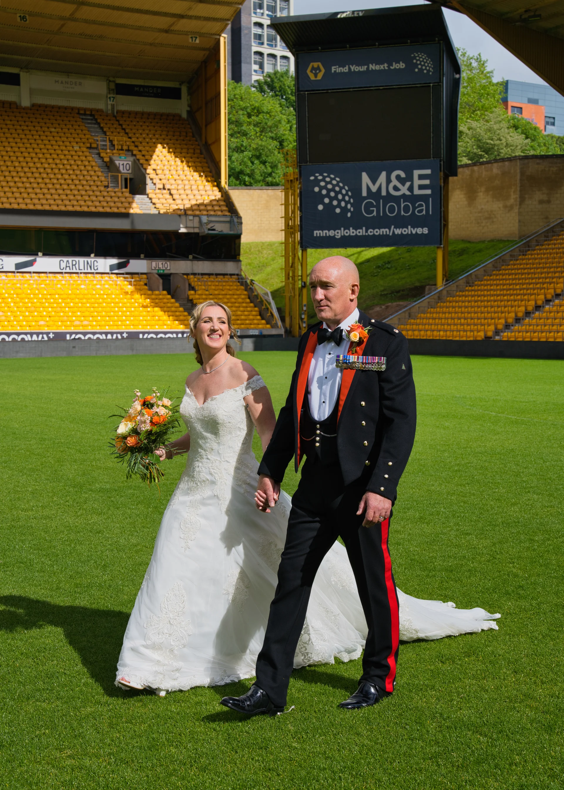 A smiling bride and a groom in a military uniform walk hand in hand across the green pitch of a large football stadium.