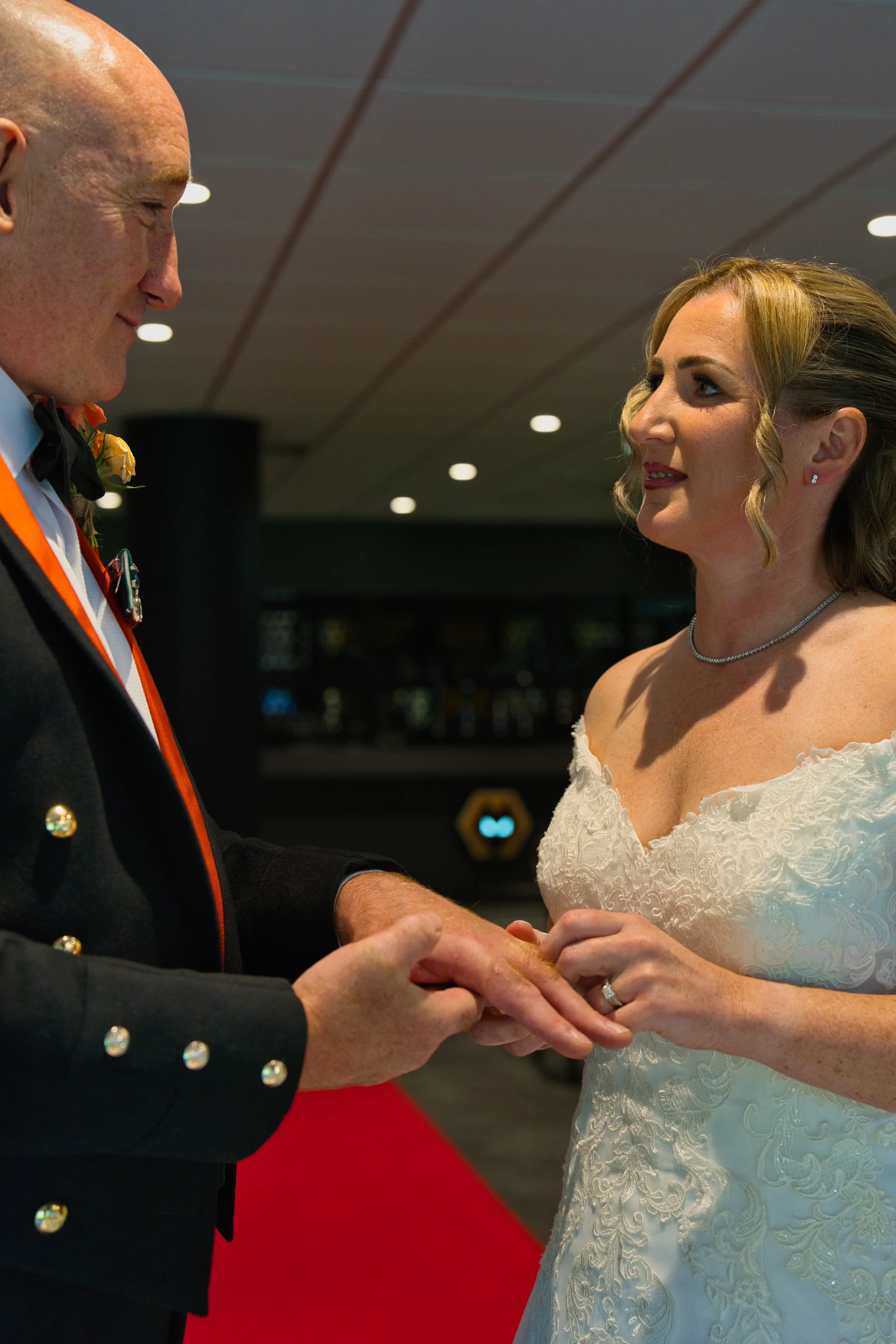 The bride and groom exchange rings during their indoor ceremony, with the bride wearing an off-the-shoulder lace dress and the groom in a military uniform.