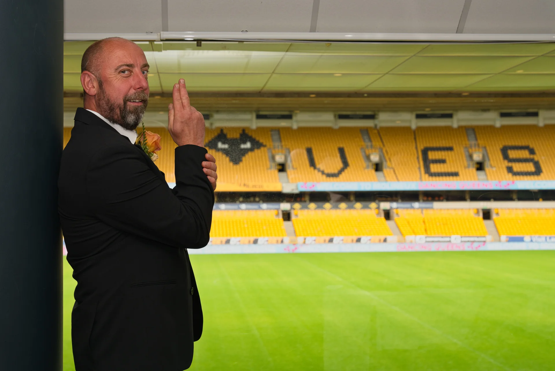 A groom in a black suit poses inside a sports stadium near seating and the pitch.