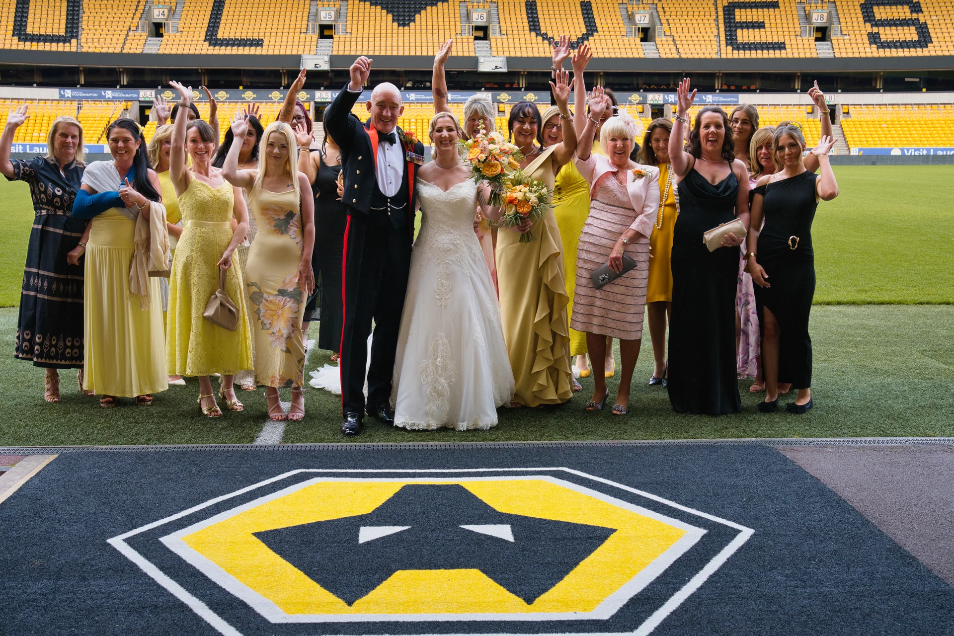 A newly married couple poses with a large group of wedding guests on the pitch inside a yellow and black football stadium, with everyone waving their hands in the air.